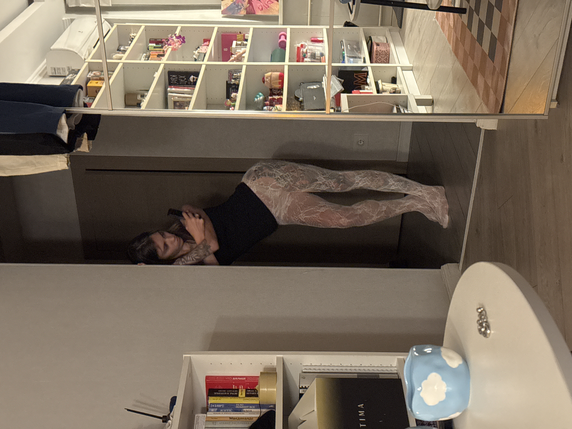 An adult female is standing in a doorway, leaning against the wall while looking at her phone. She is wearing a black tank top and white lace leggings. To her left, a white bookshelf is filled with books and decorative items. To her right, a tall white bookshelf displays various objects, and a large mirror reflects the room. The floor is made of light-colored wood. A round white table with a blue cloud-shaped ceramic dish and some small metal objects is visible in the foreground. The scene appears to be an indoor residential setting.