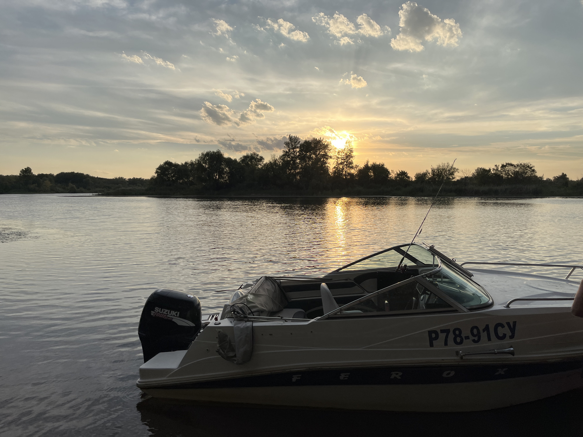 A boat is docked on a body of water next to a tree line at sunset. The sky is filled with wispy clouds, and the sun is setting behind the trees, casting a golden reflection on the water. The boat is white with a black outboard motor that has the Suzuki logo on it. The boat has a registration number that reads P78-91CY. The water appears calm with slight ripples.