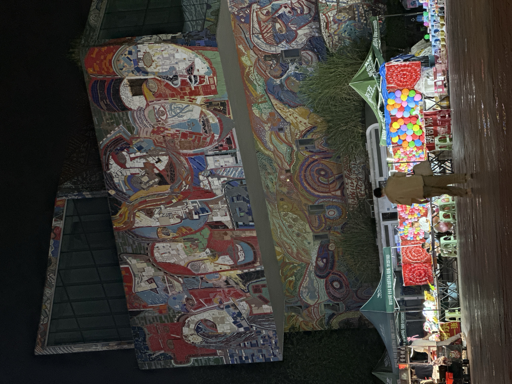 The image depicts a night scene in the Chongqing High-tech Zone, China, showcasing a lively street market situated in front of a building adorned with large, vibrant mosaics. The mosaics, featuring abstract representations of faces and colorful patterns, cover a significant portion of the building's facade. Below the mosaics, several market stalls are set up. One stall is a game booth with numerous colorful balloons and a red backdrop. Another stall appears to be selling goods from under a green tent. A few people are visible, including a person walking away from the camera, dressed in a light-colored shirt and pants, carrying a bag. Other individuals are present at the stalls, some appearing to be vendors and others customers. The ground is wet, reflecting the lights from the market, suggesting it may have recently rained. The overall atmosphere is that of a bustling night market in an urban environment.
