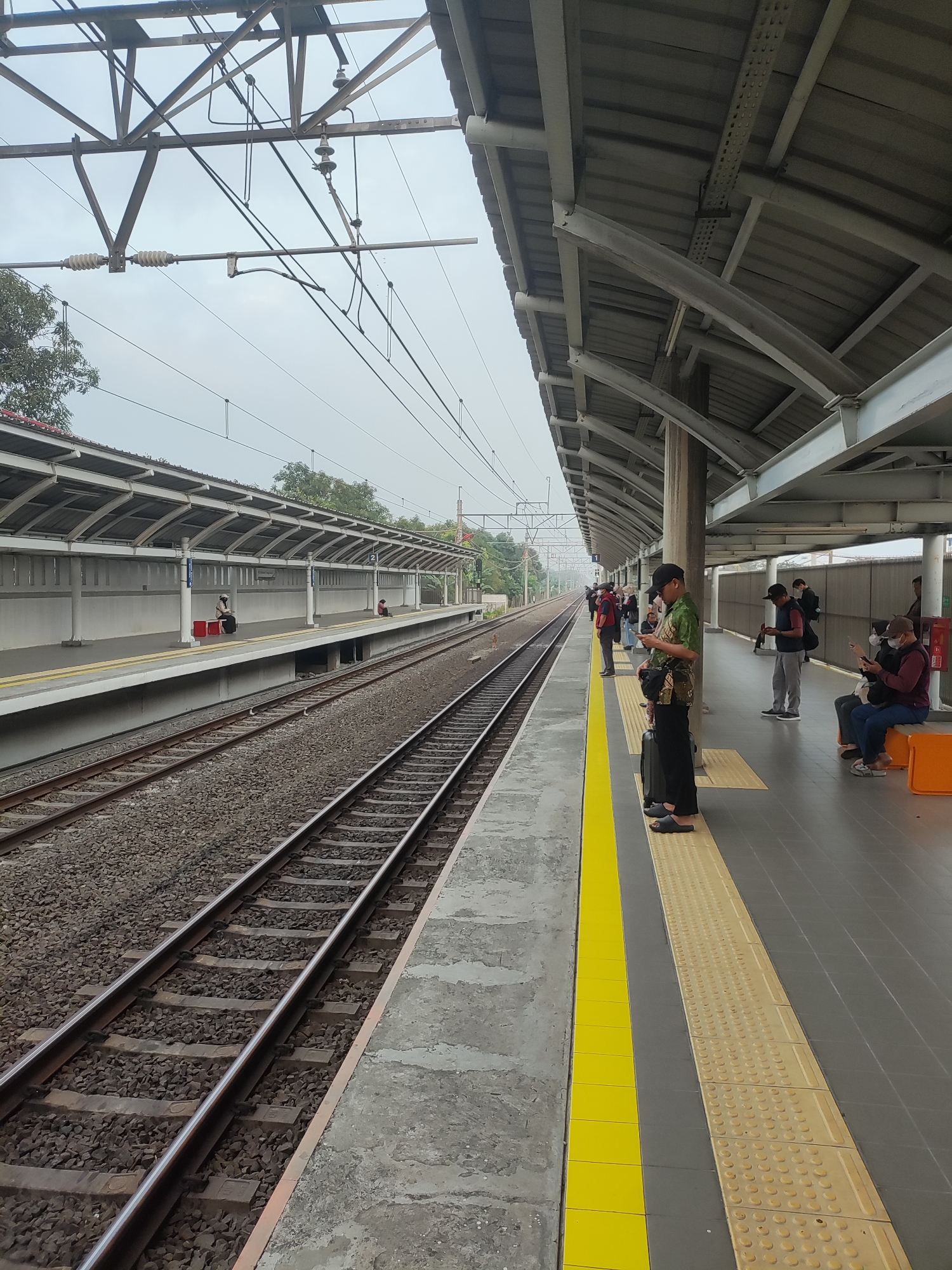 The image shows a train station platform in Telagamurni, Indonesia. The platform is covered by a corrugated metal roof supported by metal beams and columns. Railway tracks run alongside the platform, which is lined with a yellow tactile paving strip. Several people are visible on the platform, some standing and others sitting on an orange bench. They appear to be waiting for a train. The sky is overcast, and trees are visible in the background. The overall scene depicts a typical day at a suburban train station.
