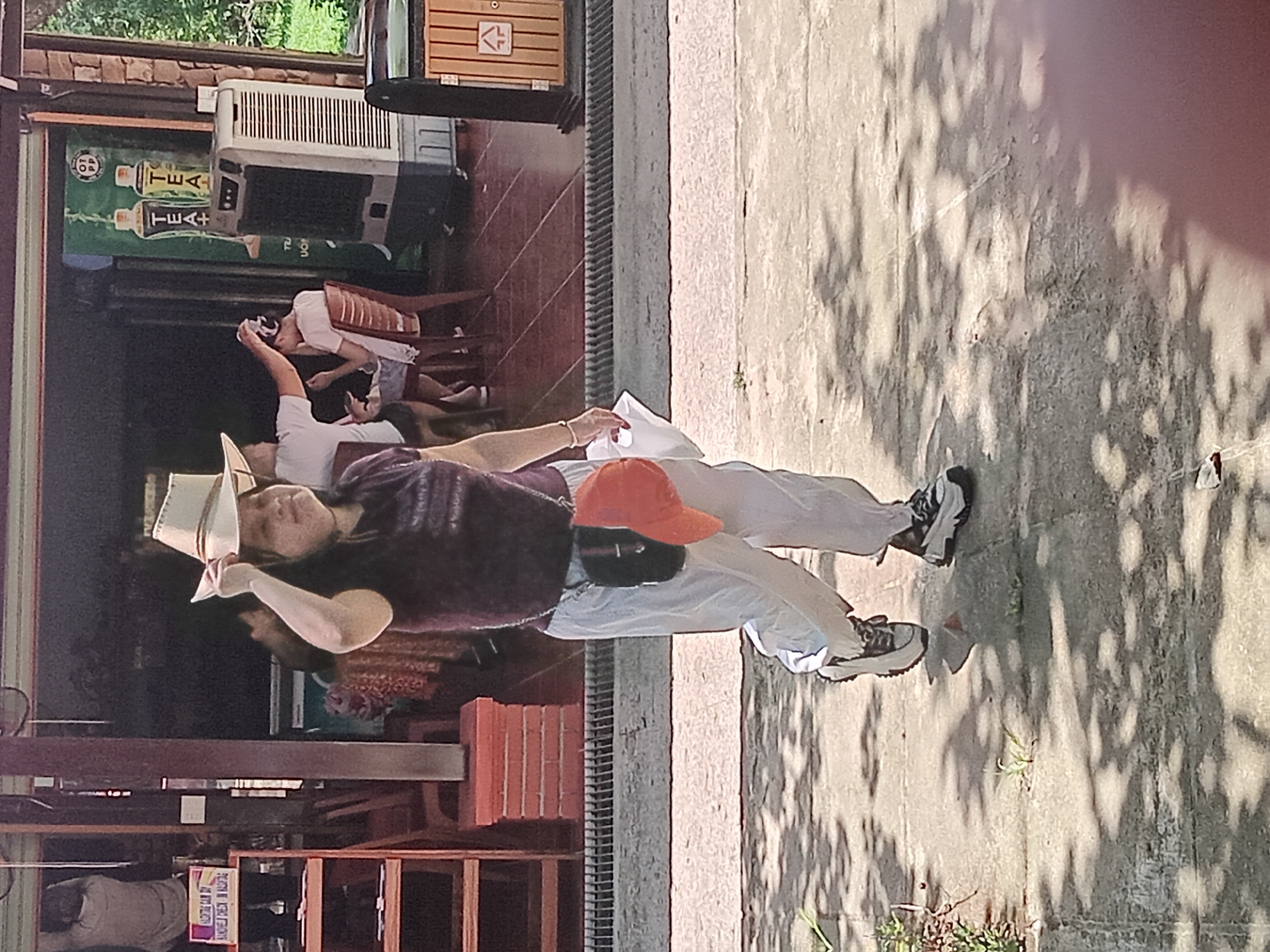 A woman wearing a cowboy hat and a purple t-shirt walks across a concrete path. She is wearing white pants and sneakers. Her right hand is raised, touching her hat. A small black bag with an orange cap is hanging from her shoulder. In the background, there is a building with a thatched roof and a refrigerator with "TEA" branding. The scene is outdoors on a sunny day, with shadows from trees cast on the ground.