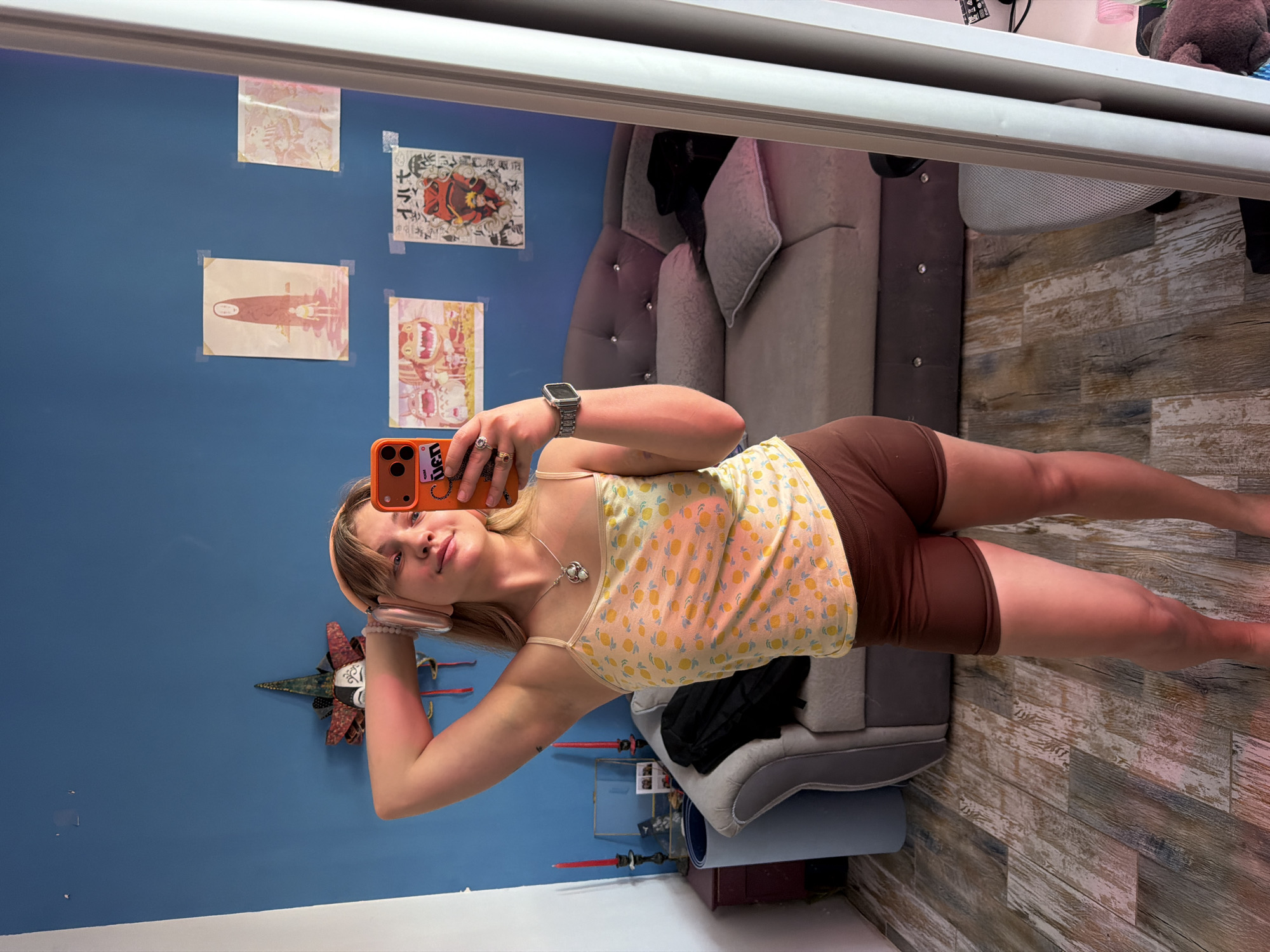 The image is a mirrored selfie taken indoors. A young woman with blonde hair and fair skin is holding an orange smartphone, presumably taking the picture. She is wearing a yellow tank top with a lemon print, and brown shorts. She also has a pink headband and a silver necklace. A watch is visible on her left wrist.
Behind her is a blue wall with several anime-style posters taped to it. To the right of the woman, there is a grey cushioned sofa/bed with a black garment draped over it. A light grey office chair is visible next to the sofa. The floor appears to be made of light brown planks with a wood grain pattern.
The location is unspecified but may be in Большая Шильна, Russia.