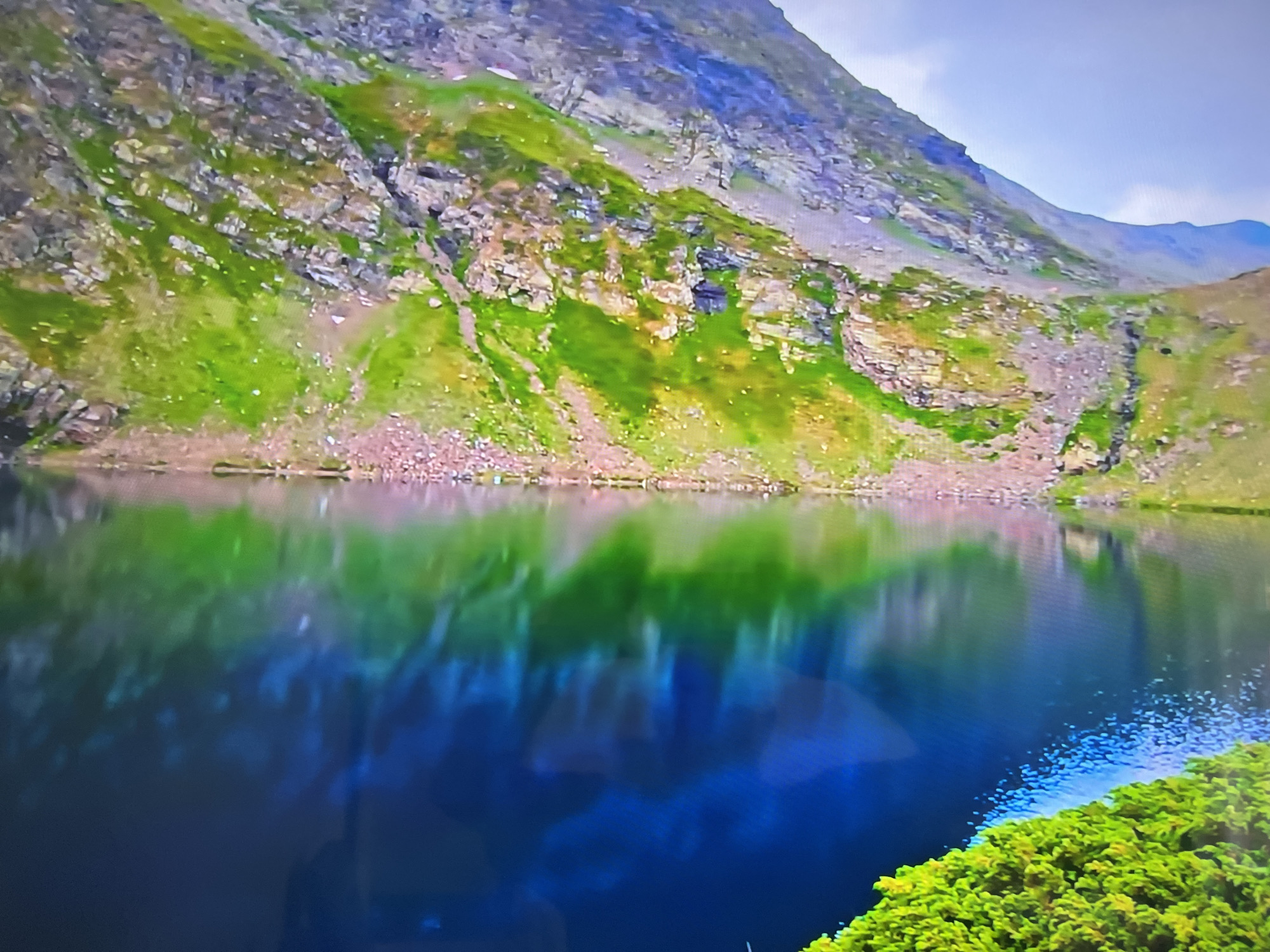The image depicts a mountain lake scene. The lake is deep blue with reflections of the surrounding green and rocky terrain. A mountain rises sharply behind the lake, covered with green vegetation interspersed with rocks. The location is Unknown, Unknown.