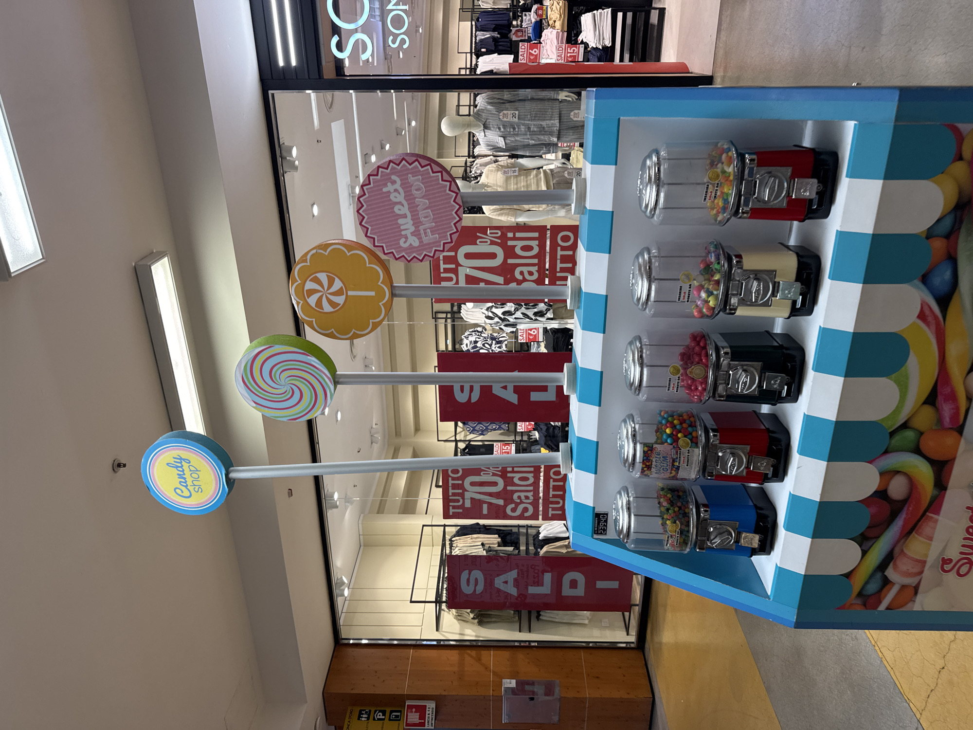 The image depicts a candy shop display within a shopping mall. In the foreground, a brightly colored stand showcases four gumball machines filled with colorful gumballs. The stand itself is decorated with a blue and white striped awning pattern and a graphic of candy canes and lollipops on its lower front. Above the stand, large, stylized lollipop signs on poles are displayed, including one reading "Candy shop" and another with a swirling pattern. In the background, the storefront of a clothing store is visible, featuring mannequins and racks of clothing with large "SALDI" (sale) signs indicating discounts. The overall environment appears to be an indoor shopping center. The image was taken in Palermo, Italy.