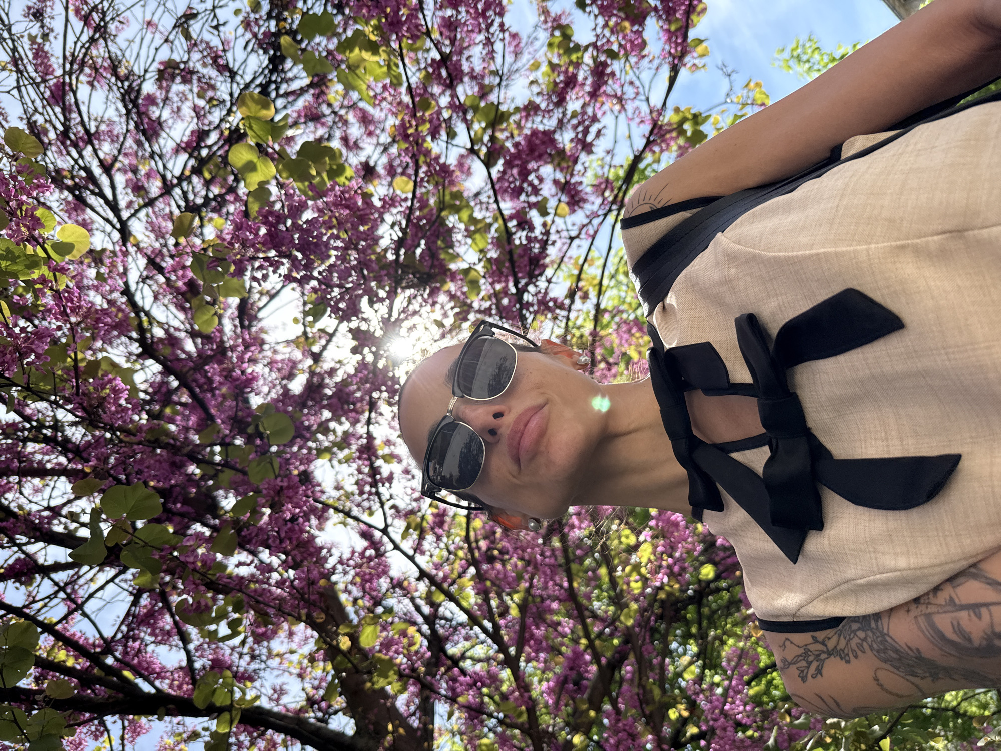 This image presents a unique, low-angle perspective of a person posing under a blossoming tree. The subject, visible from the chin upwards and angled downwards into the frame, wears sunglasses and a light-colored top with distinct black ribbon-like detailing and bows. A tattoo, resembling a stylized sun or radiating lines, is visible on their right shoulder, and another intricate, possibly floral, design adorns their lower right arm. The person exhibits a subtle, relaxed smile.
The backdrop is dominated by a tree in full bloom, featuring abundant clusters of vibrant purple or magenta flowers and scattered green leaves, typical of a Redbud tree. Bright sunlight filters through the branches, creating a lens flare and illuminating the scene. Patches of clear blue sky are visible between the dense foliage and blossoms. The overall impression is a cheerful, sunny spring day. The location context for this vibrant outdoor scene is Pleven, Bulgaria. No discernible text is visible.