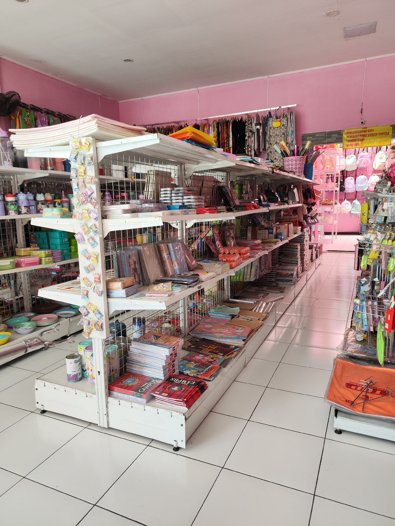 The image depicts the interior of a retail store selling stationery, school supplies, and children's items. The store is characterized by its pink walls and white shelving units filled with a variety of merchandise. On the shelves, one can observe stacks of notebooks, folders, pens, rulers, craft supplies, and decorative items. Several backpacks are displayed on a rack in the background, along with hanging clothing items. The floor is tiled with white squares and black grout lines. A yellow sign with text in Indonesian is visible on the back wall, indicating that this store is located in Sragen, Indonesia. The scene is well-lit, suggesting it is daytime. There are no people visible in the image.
