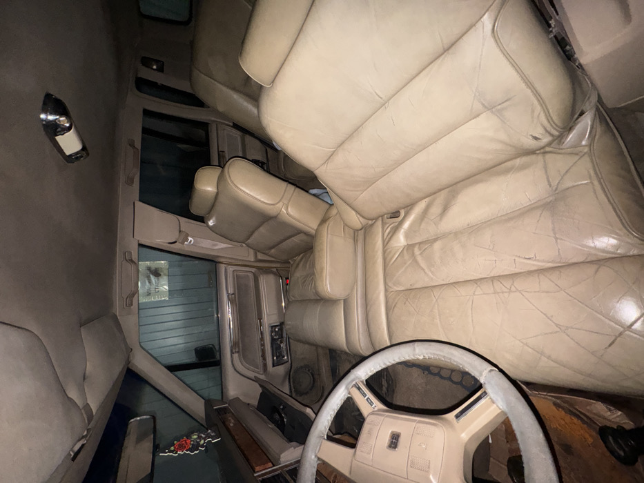 The image displays the interior of an unoccupied light brown/beige vehicle, identified as being in Zhabkino, Russia, based on external cues. The perspective is from the driver's side floor, looking across the front passenger area towards the rear seats.
**Subjects and Objects:**
The primary subject is the vehicle's interior. A beige four-spoke steering wheel, showing wear on its rim, is partially visible in the lower left foreground. The seating consists of worn, tan leather or faux leather upholstery, exhibiting numerous creases, cracks, and some discoloration across all visible seats. The front passenger seat is reclined. A dashboard with a dark upper surface and a prominent wooden trim accent is visible on the passenger side. A black rose-shaped air freshener hangs from the rearview mirror, which is partially visible. An overhead dome light fixture with a chrome base is mounted on the vehicle's ceiling. Door panels, matching the interior color, are visible, featuring power window controls.
**Environment:**
Through the vehicle's side and rear windows, a light grey-green corrugated metal wall is visible, indicating the vehicle is parked inside a garage, storage unit, or similar enclosed structure. A white piece of paper with dark Cyrillic text is taped to this external wall, visible through the front passenger window, confirming a Russian setting. The interior is dimly lit, suggesting low ambient light conditions.
**People, Actions, and Interactions:**
No people are present in the image. No actions or interactions are depicted. The scene is static.