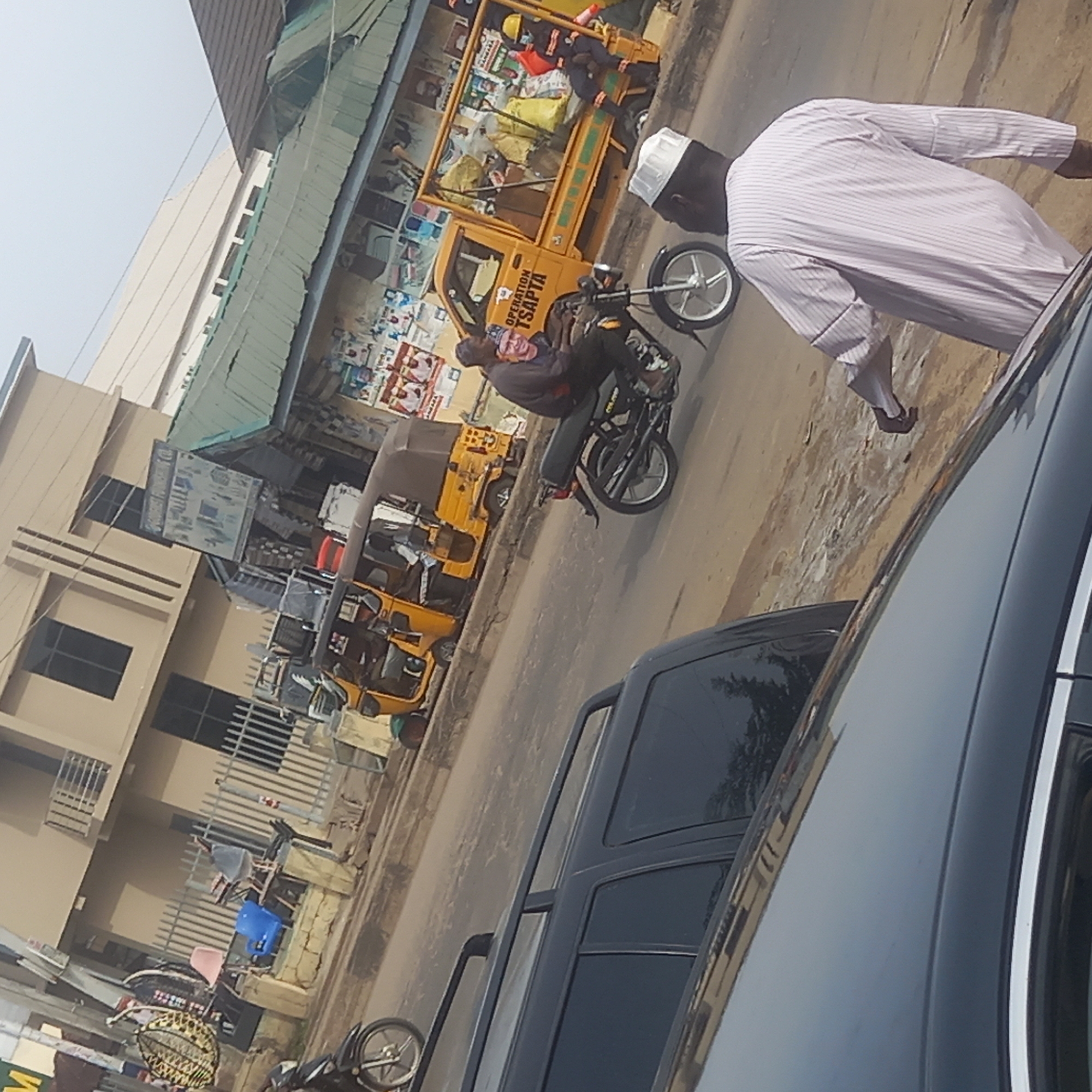 The image shows a street scene in what appears to be Dadi Riba, Nigeria. A yellow "Keke Napep" tricycle taxi labeled "OPERATION TSAPTA" is parked on the side of the road. A man wearing a white skullcap and a striped shirt is walking near the vehicle. A motorcycle with a rider is also present. Buildings and shops are visible in the background, along with various items stacked near the shops. A black vehicle is partially visible in the foreground.