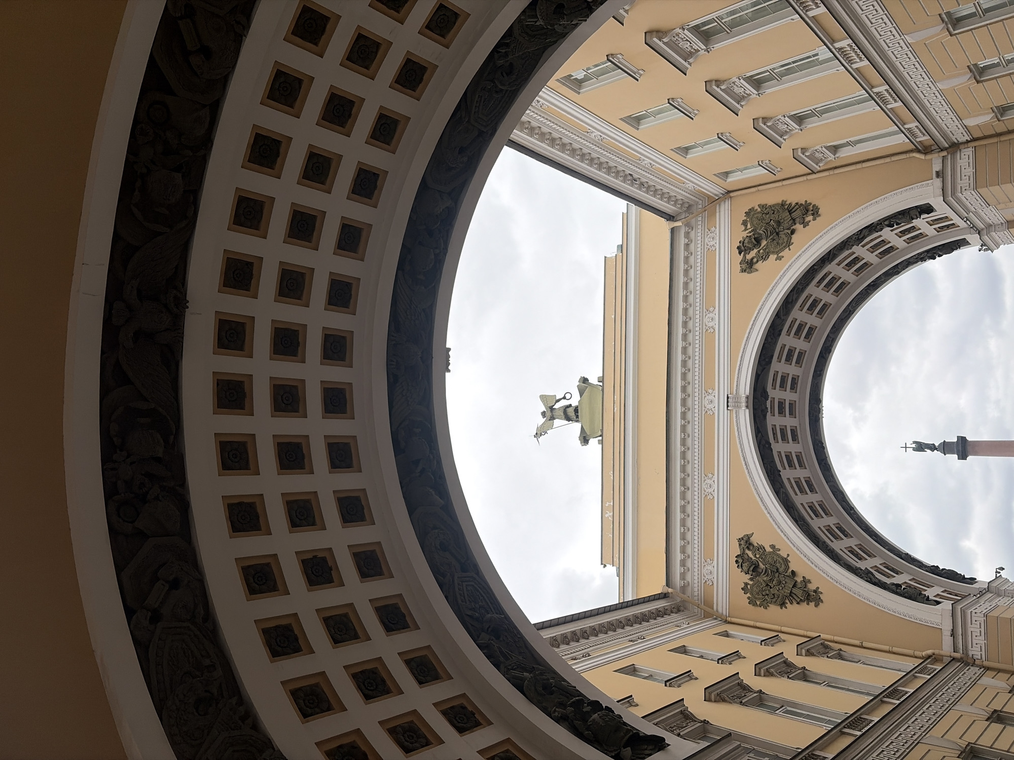 The image presents a dramatic, upward-looking perspective through a monumental archway, showcasing the opulent classical architecture of Saint Petersburg, Russia. The arch's underside features a grid of square coffers, each detailed with a dark, circular rosette motif. Along the inner curve of the arch, elaborate bas-relief carvings depict figures, winged mythological creatures, and intricate scrollwork, all rendered in dark stone against a lighter backdrop.
Beyond the archway, pale yellow buildings with multiple rectangular windows and white trim rise towards the overcast sky. These structures are richly adorned with neoclassical elements, including detailed cornices, decorative friezes (some featuring Greek key patterns), and ornate wall panels, possibly gilded cartouches with military or imperial emblems.
Dominating the view above the buildings, and framed by the arch, is a large sculptural group, likely depicting a triumphant figure, possibly in a chariot or holding a spear, positioned atop a structure. In the far distance, the iconic Alexander Column is clearly visible against the cloudy sky, distinguished by its tall, slender shaft and the statue of an angel holding a cross at its summit. The scene captures the grandeur and historical significance of Palace Square, with the Arch of the General Staff Building framing the Alexander Column. No specific activity or visible text is present; the image is a static architectural study on a cloudy day.