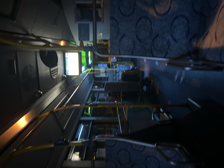 The interior of a bus is captured in a blurry image. Rows of seats with blue patterned upholstery stretch down the aisle. Yellow handrails run along the ceiling. A digital display screen glows near the front. Windows line the sides, and luggage racks are visible above the seats. The overall lighting is dim.