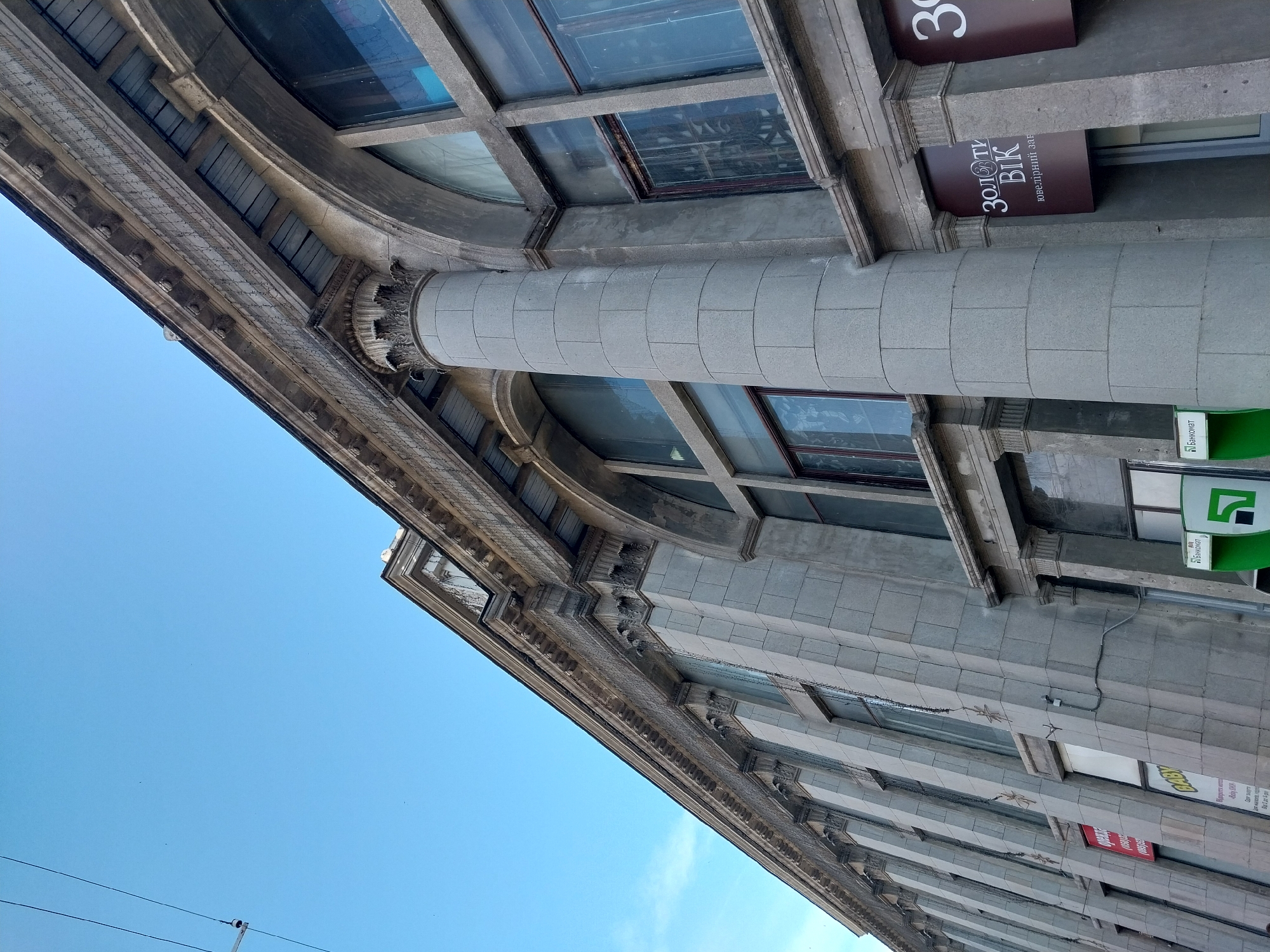 The image shows the exterior of a building with classical architectural elements, including columns and arched windows. The building appears to be made of stone or concrete blocks. There are signs on the building, some of which are in Ukrainian. One sign reads "ЗОЛОТИЙ ВІК" and another has the word "БАНКОМАТ" (ATM). The sky is clear and blue. The perspective is a low-angle shot looking up at the building. This is a view of the building at 31 Velyka Dymerka Street in Dnipro, Ukraine.
