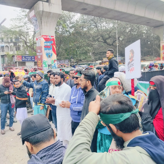 A crowd of people are gathered outside in front of a building and under a bridge. The group appears to be engaged in a protest or demonstration, as one person is holding a sign that says "Justice for Hadi" along with a portrait. The people are diverse in age, with some wearing traditional Muslim clothing and others in modern attire. The atmosphere seems serious and determined.