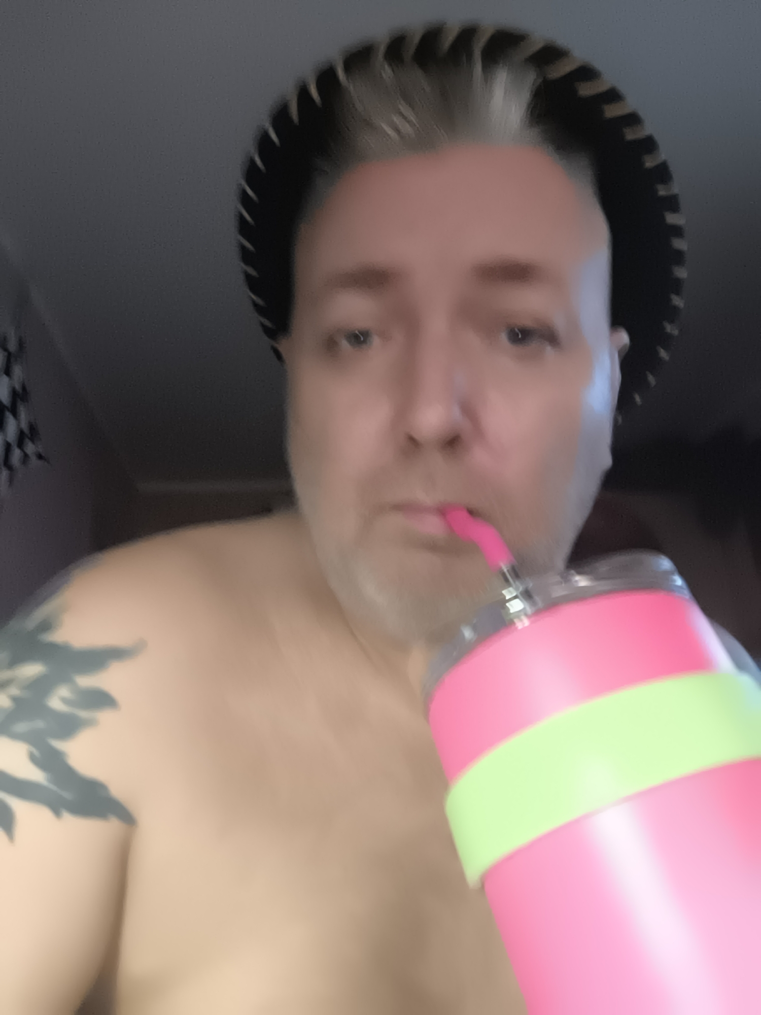 The image shows a close-up, slightly blurry, selfie of a person with fair skin, light-colored hair, and a short beard. They are wearing a black and brown patterned hat that resembles a sombrero and have a tattoo on their left shoulder and upper arm. The person is holding a pink and light green tumbler with a pink straw to their lips, appearing to drink from it. The background is dark and indistinct, with a hint of a checkered pattern visible on the left side. The scene is captured indoors, and no specific location in Riga, Latvia, can be identified from the visual information provided.