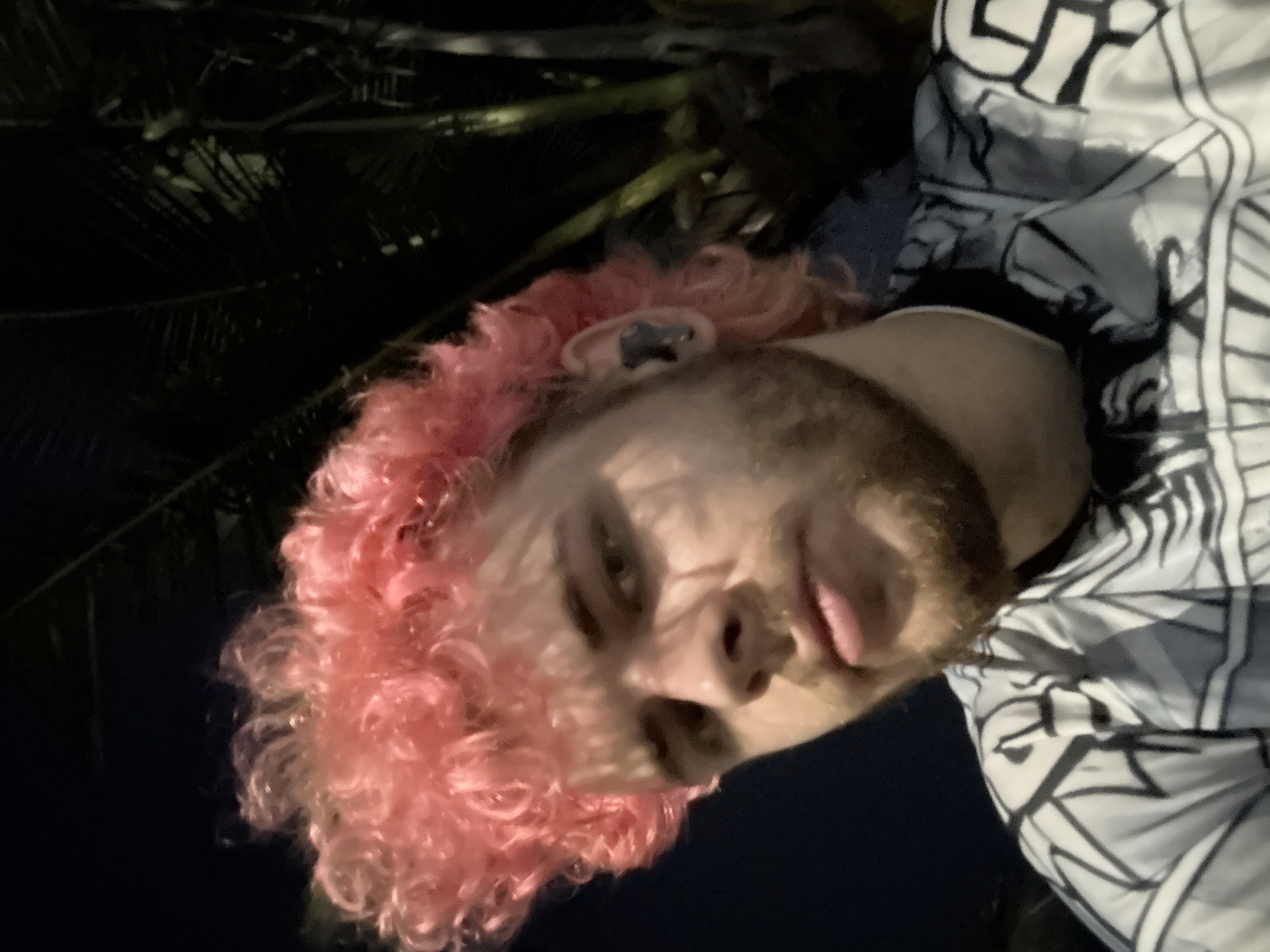 A person with pink curly hair and a beard is facing the camera, with a dark background of palm leaves. The person is wearing a patterned black and white shirt. The location cannot be determined to be in Đà Nẵng, Vietnam with the given information.