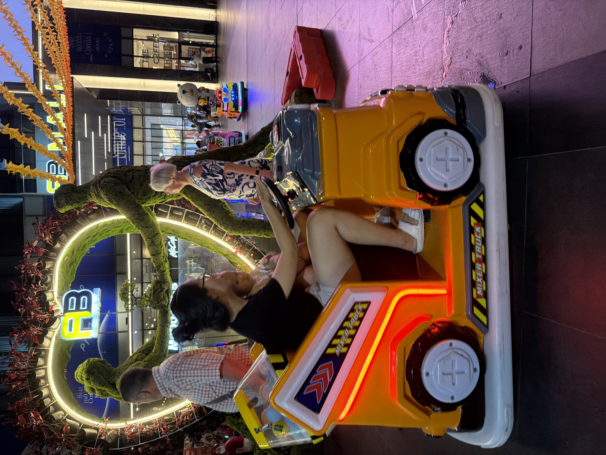 A woman and a baby are seated in an orange toy mixer truck, a type of amusement ride. The woman is steering the truck, while the baby sits in her lap. The truck is positioned on a tiled ground surface illuminated by purple lighting. In the background, several people are visible, and a large heart-shaped structure adorned with greenery and neon lights stands prominently. A sign with the text "AB SHOPPING MALL" is displayed on the structure. The overall environment suggests an outdoor setting, possibly a mall plaza or entertainment area in Nha Trang, Vietnam.