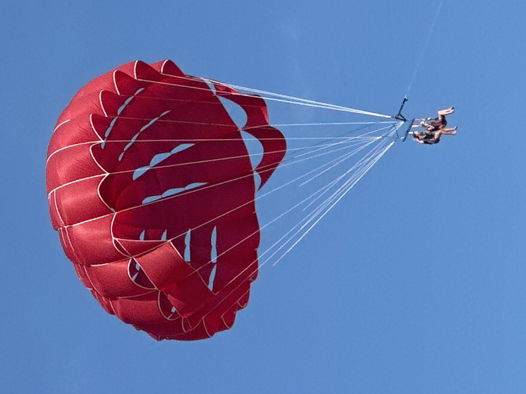 A red parasail with white trim is open and aloft against a clear blue sky. The parasail is angled slightly to the right. Below the parasail, two individuals are suspended in a harness, appearing to be parasailing. The figures are small in the frame. The image is a close-up, likely taken from a distance or a lower angle. The location is not identifiable from the image, and there is no indication of Boreti, Montenegro.