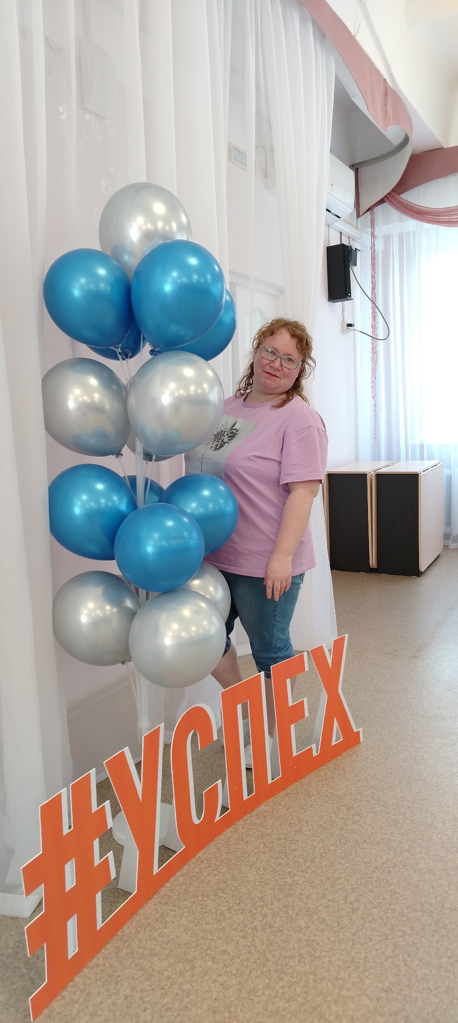 A woman stands in a room with blue and silver balloons near a sign that reads “#УСПЕХ” (which translates to #Success). The woman has red curly hair, wears glasses, a pink t-shirt with a print on it, and blue jeans shorts. The balloons are arranged in a cluster and are mounted on a stand. The sign is orange with white lettering. The location appears to be inside a building in Chaikovsky, Russia, based on the Russian text on the sign. In the background, there is a white curtain, an air conditioning unit, a speaker mounted on the wall, and cabinets. The floor is light brown.