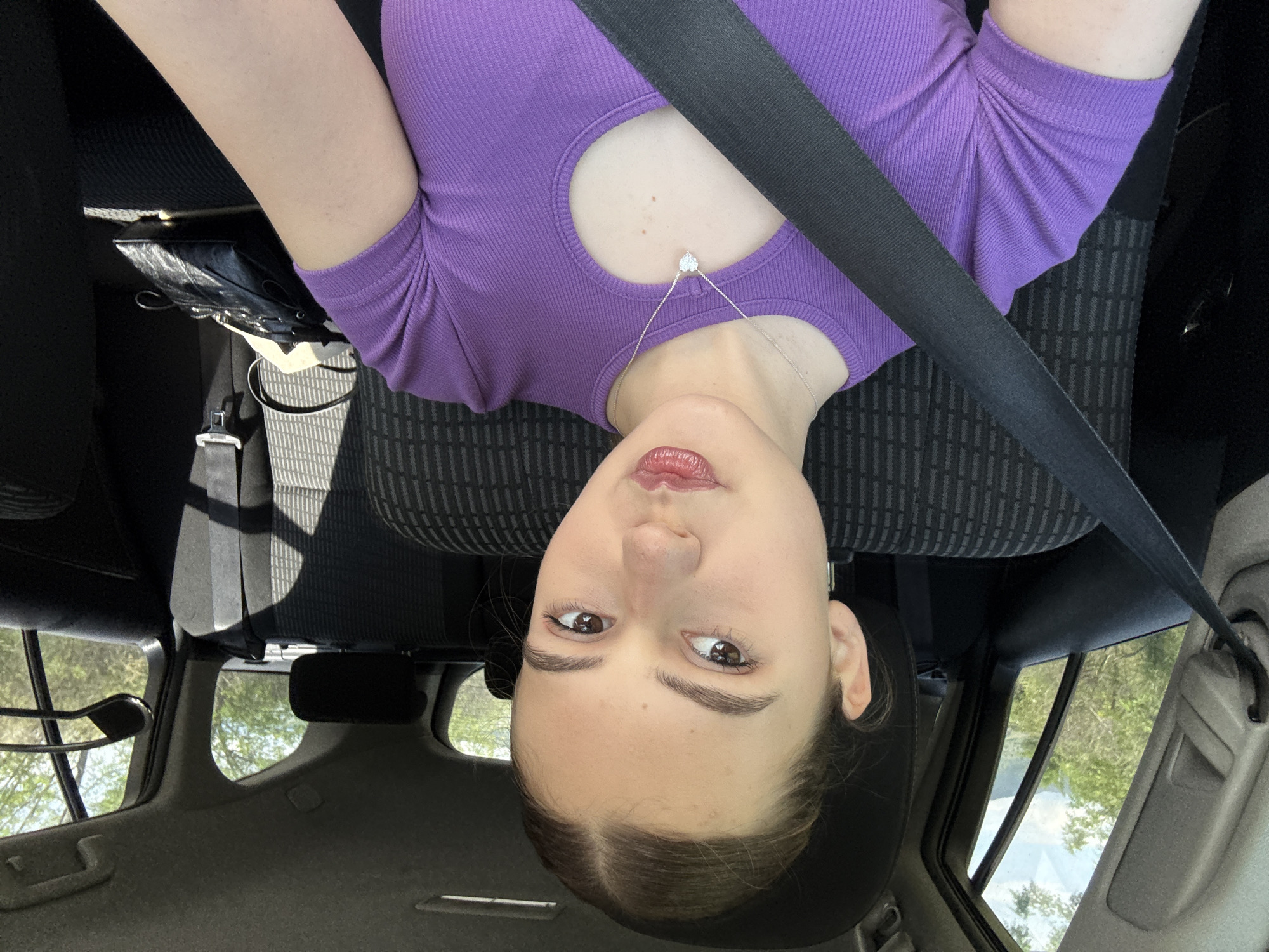 A fair-skinned young woman with brown hair pulled back is shown from an upside-down perspective in the interior of a car. She is wearing a purple, ribbed shirt with a circular cutout below the neck and a silver necklace with a small pendant. She is wearing a black seatbelt diagonally across her torso. The car's seat is upholstered in a patterned fabric. The interior of the car's roof, windows, headrest, and dashboard are visible. Through the car window, trees and sunlight can be seen. The city of Levski, Bulgaria is not identifiable from the image.