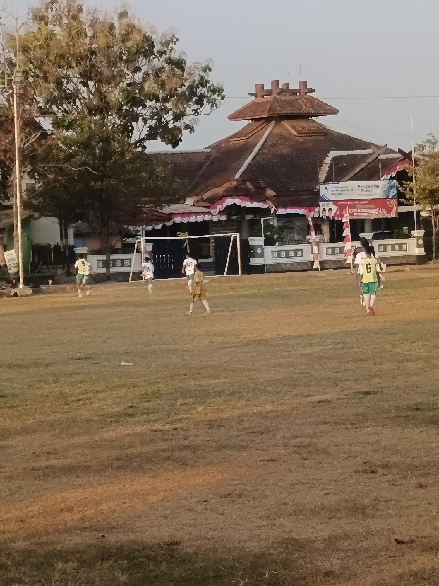The image depicts a soccer game taking place on a dry, grassy field in front of a building with traditional Indonesian architecture. Several young males, dressed in soccer attire, are visible on the field. One player, wearing a yellow jersey with the number 9, is in possession of the ball. Another player in a white jersey with the number 5 is running towards the goal. A third player, in a white jersey with the number 47, is standing near the goal. A fourth player, wearing a yellow and green jersey with the number 6, is walking away from the camera. The building in the background has a distinctive multi-tiered roof and is adorned with decorative banners. The overall scene suggests a casual, outdoor sporting event in a village setting.