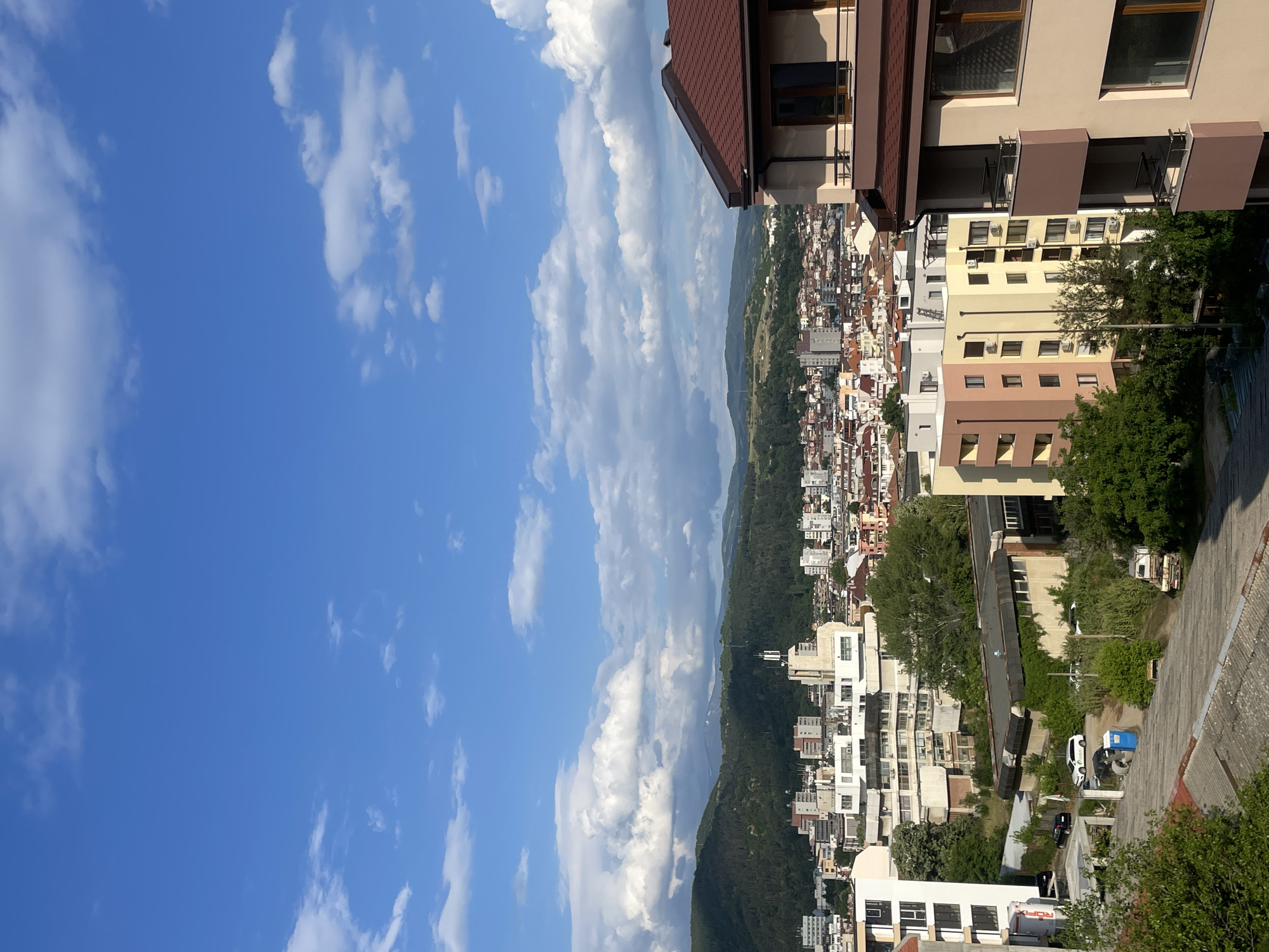 A panoramic view captures a sun-drenched cityscape nestled against a backdrop of green hills, all under a vast blue sky adorned with scattered white clouds. The image is taken from an elevated perspective, likely from an upper floor or rooftop of a building.
In the immediate right foreground, part of a modern building is visible, featuring a prominent reddish-brown tiled roof and light beige facades. Several rectangular windows with dark frames and balconies with dark railings can be seen on this structure.
The midground and background showcase a sprawling urban area, extending from the center of the frame towards the left and into the distance. This part of the city is densely populated with numerous multi-story residential and commercial buildings, displaying a variety of colors including light yellow, beige, brown, and white. The architecture suggests a mix of contemporary and slightly older European styles. Green trees and other vegetation are interspersed throughout the urban fabric, breaking up the concrete landscape and adding natural elements.
Beyond the city, rolling green hills and mountains rise, forming a natural boundary and backdrop, indicating the city is situated in a valley or mountainous region.
The sky is a brilliant, clear blue, filled with scattered, fluffy cumulus clouds, characteristic of good weather. The strong sunlight casts distinct shadows, suggesting the time of day is likely late morning or early afternoon.
No specific human activity is evident in the broad shot. However, closer examination of crops reveals a white delivery truck, a white car, a black car, and a blue trash bin on what appears to be a street in the lower parts of the city. On one white cylindrical tank attached to a building, the text "NORAX" is visible. The scene is consistent with the urban landscape of Blagoevgrad, Bulgaria, known for its mountainous surroundings.