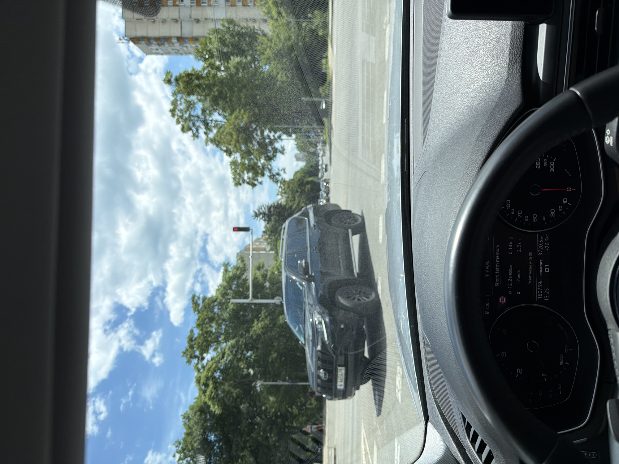 The image is taken from the interior of a car, looking through the windshield. A black Toyota Land Cruiser Prado is visible in the foreground, stopped at an intersection. The car has a license plate that appears to be A 6656 PH. The intersection shows traffic lights, road markings, and other vehicles in the distance. Trees line the sides of the road, and a tall apartment building is visible in the background. The sky is blue with scattered white clouds. On the car's dashboard, a digital display shows information including speed, fuel consumption, and time. The location is specified as Burgas, Bulgaria. The scene appears to be a typical urban street on a sunny day.