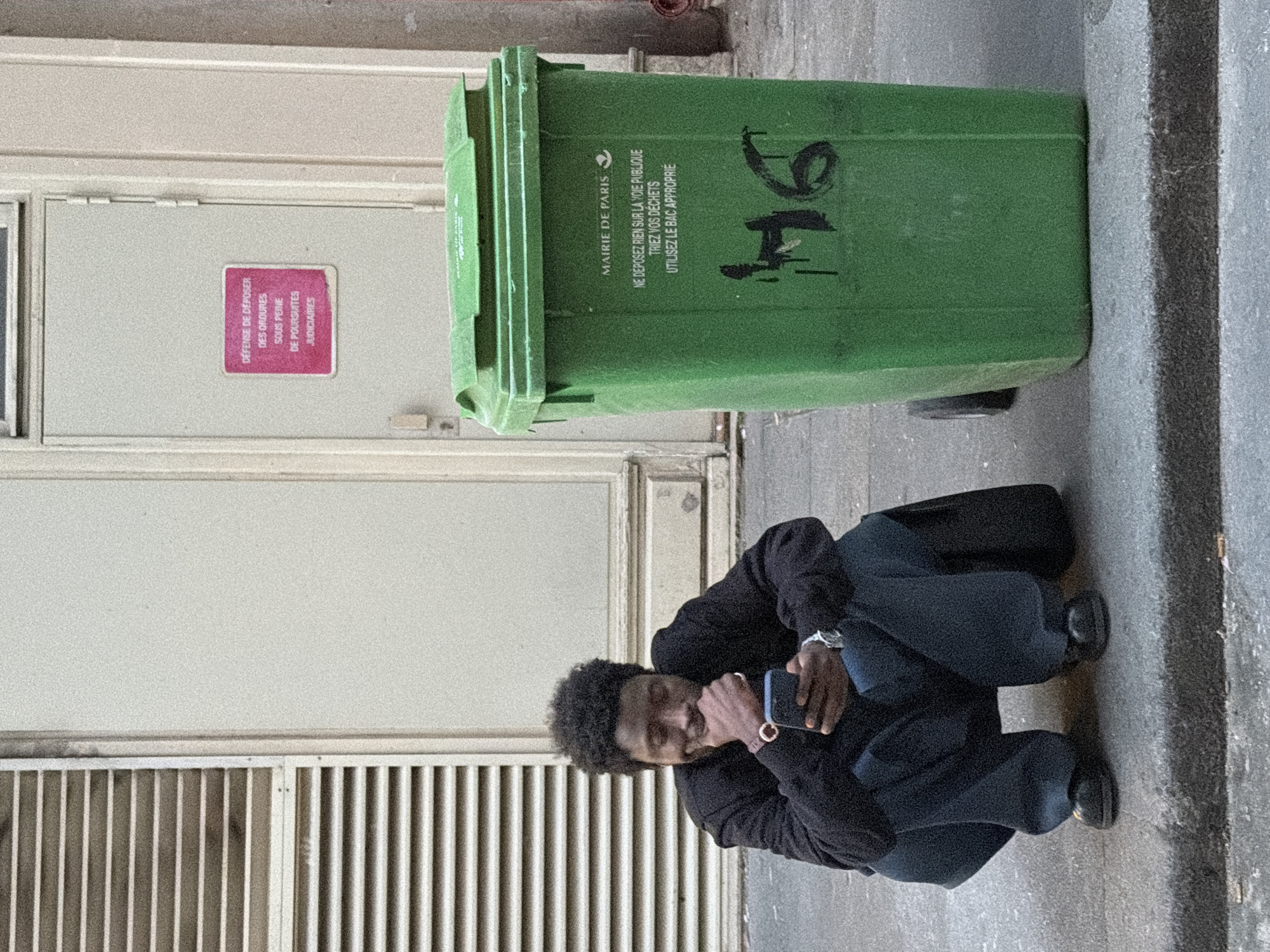 The image shows a man crouching next to a green trash bin in an urban environment, likely Paris, France. The trash bin has the words "MAIRIE DE PARIS" (City Hall of Paris) printed on it, along with instructions to not place anything on the public road and to sort waste using the appropriate bin. The number "94" is spray-painted on the bin. The man is looking at a phone and wearing a dark jacket and dark pants. Next to the trash bin and man is a door with a pink sign that reads "DÉFENSE DE DÉPOSER DES OROURES SOUS PEINE DE POURSUITES JUDICIAIRES." The door has a grate at the bottom. The street or sidewalk appears to be concrete.