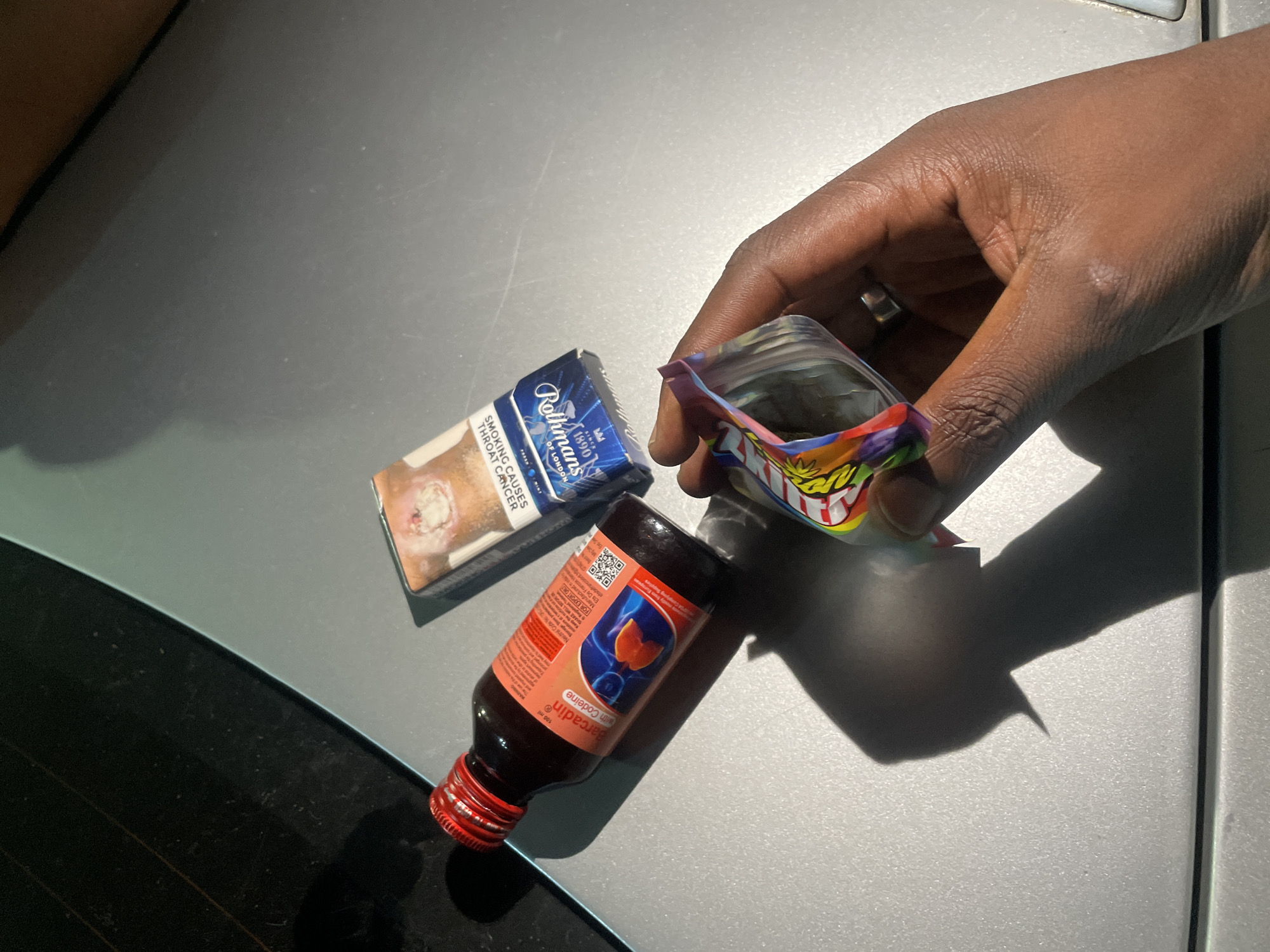 A close-up, high-angle image depicts a dark-skinned hand holding a small, colorful plastic bag, positioned against a bright, reflective surface, likely the hood or trunk of a car. The hand, wearing a ring on its index finger, is opening the bag, revealing green, leafy plant matter inside.
On the same reflective surface, two other items are prominently displayed. A packet of Rothmans cigarettes, characterized by its blue and white packaging, rests near the hand. Visible text on the cigarette packet includes "Rothmans," "SINCE 1890," "OF LONDON," and the health warning "SMOKING CAUSES THROAT CANCER." Adjacent to the cigarettes and below the hand is a small, dark brown bottle with a red screw cap. The label clearly identifies it as "Barcadin with Codeine," a medicinal syrup. Further text on the bottle includes "100 ml," "FOR EXPORT ONLY," "Manufactured mostly from European Complying Supplies," "Ets De Francis," and a QR code.
The setting appears to be outdoors in bright daylight, evidenced by the strong shadows cast by the hand and objects. The smooth, grey surface reflects sunlight, indicating an outdoor environment, possibly on a vehicle. The background shows blurred details of a car's window or interior. The arrangement of cigarettes, codeine syrup, and the contents of the plastic bag suggests a context related to recreational drug use. The scene is located in Abuja, Nigeria.