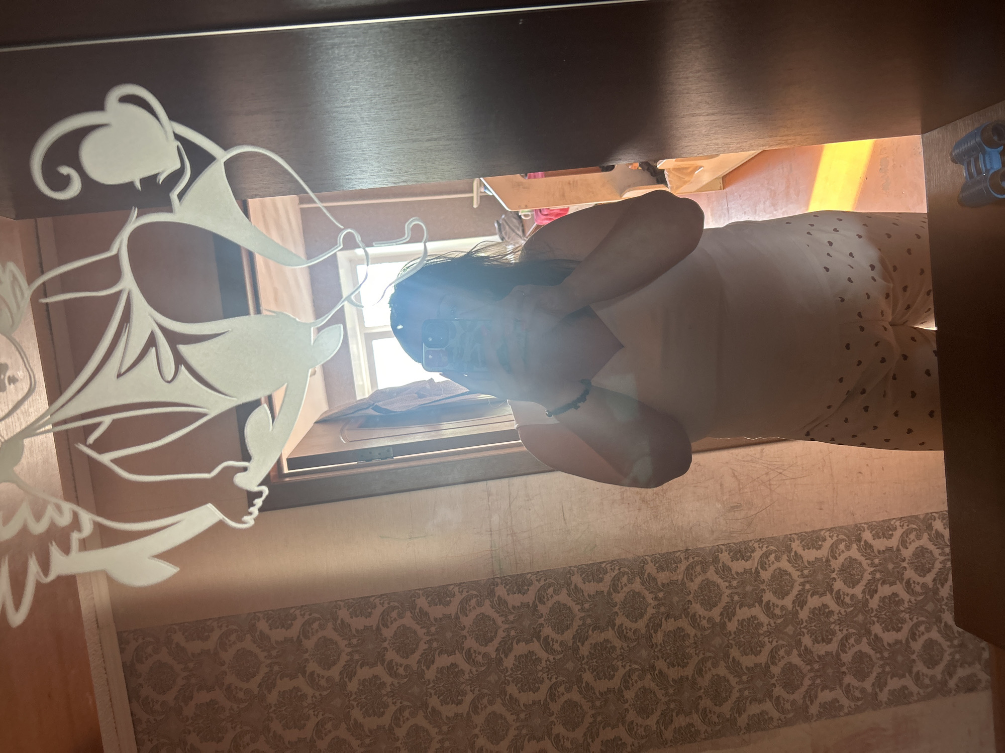 The image shows a person taking a selfie in a mirror. The person is wearing a light-colored tank top and patterned shorts. The mirror has a decorative etching of two figures. The background appears to be a room with patterned wallpaper and a doorway leading to another area where sunlight is casting a bright orange hue on the floor. There is a pair of blue binoculars on a surface in the foreground. The image does not contain any specific landmarks or details that would allow for identification of the location within the city of Omsk, Russia.