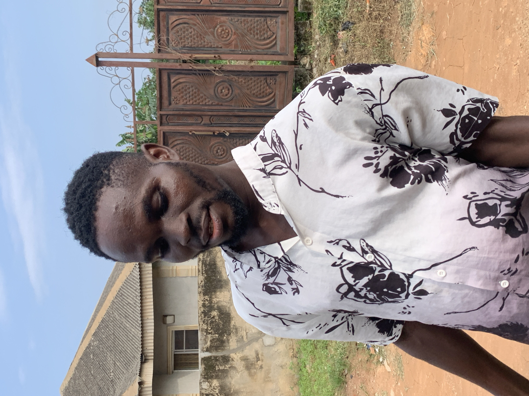 A young Black man with short, dark hair and a beard is standing outdoors, looking downwards and to his left. He is wearing a white short-sleeved shirt with a black floral pattern. Behind him, a light-colored building with a grey corrugated roof is visible to the left, and a large, ornate metal gate with intricate scrollwork and geometric patterns is behind him to the right. The ground is a reddish-brown dirt path with patches of green grass. The sky is clear and blue. The scene appears to be in Ikorodu, Nigeria, based on the architectural style and vegetation.