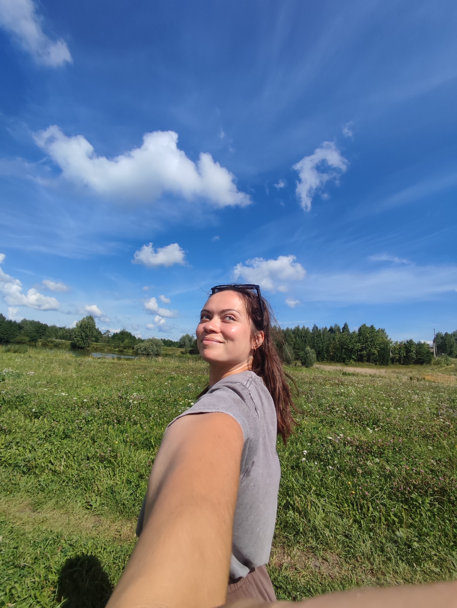 A young woman with long brown hair and sunglasses perched on her head is holding the camera out in front of her as if taking a selfie. She is wearing a grey t-shirt and a light brown skirt. She is smiling and looking up towards the sky, which is a vibrant blue with scattered white clouds. The background consists of a lush green field with various wildflowers, a body of water, and a line of trees. The overall scene suggests a bright, sunny day in a natural, rural setting. The location is specified as Fokino, Russia.