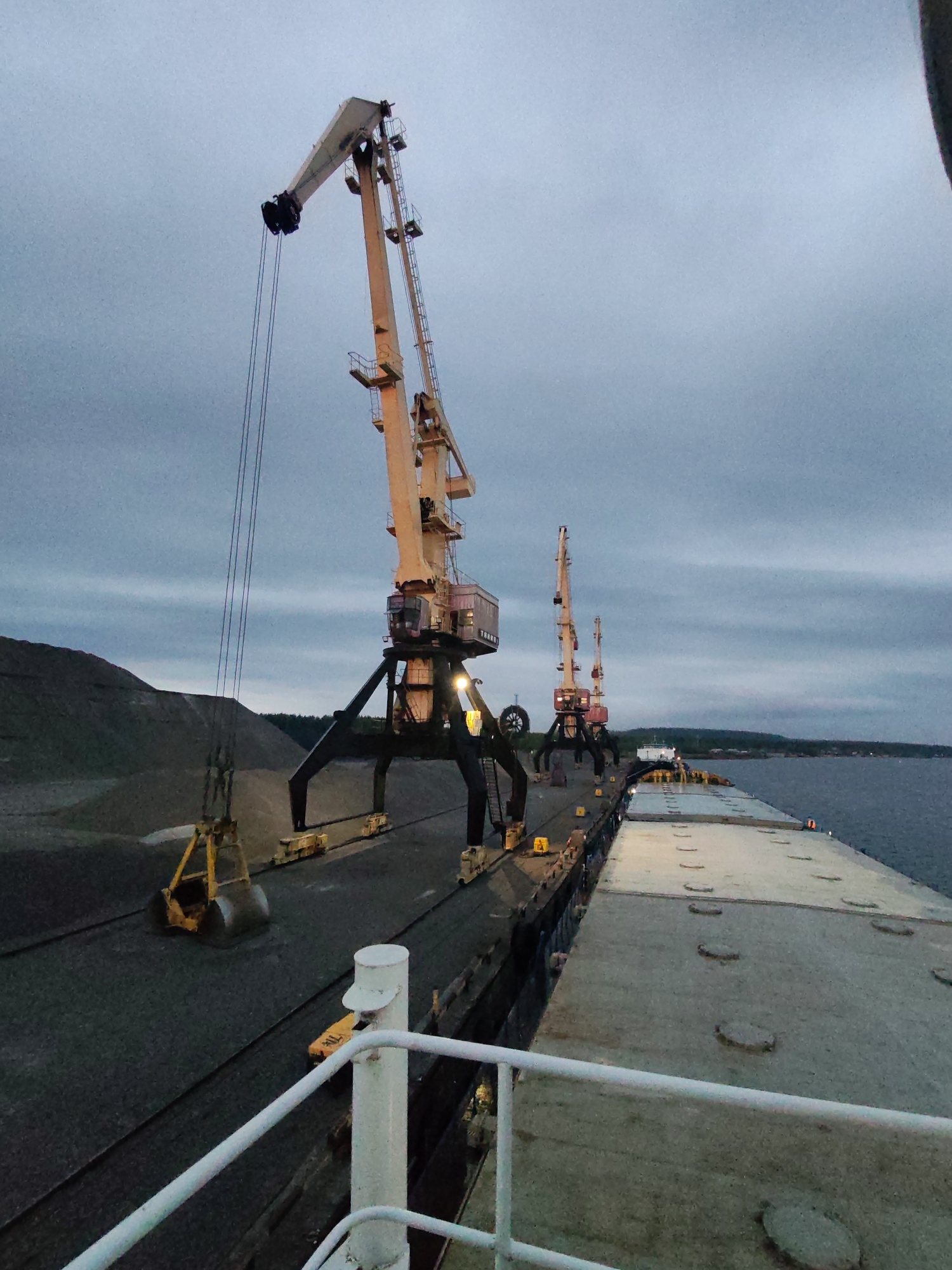 The image shows a cargo ship being loaded at a port. The scene is dominated by three large, yellow industrial cranes positioned on the dock alongside the ship. The cranes are lifting materials, presumably ore or other bulk cargo, onto the ship. The first crane is in the middle of filling up the scoop of the crane while a load of cargo, resembling gravel or ore, forms a large pile. The ship's deck is visible, constructed of rectangular sections. The sky is overcast, suggesting twilight or early morning. The presence of evergreen trees on the opposite shore suggests a coastal or inland waterway location. There is no indication of the city of Shalsky or other information that can confirm its location.