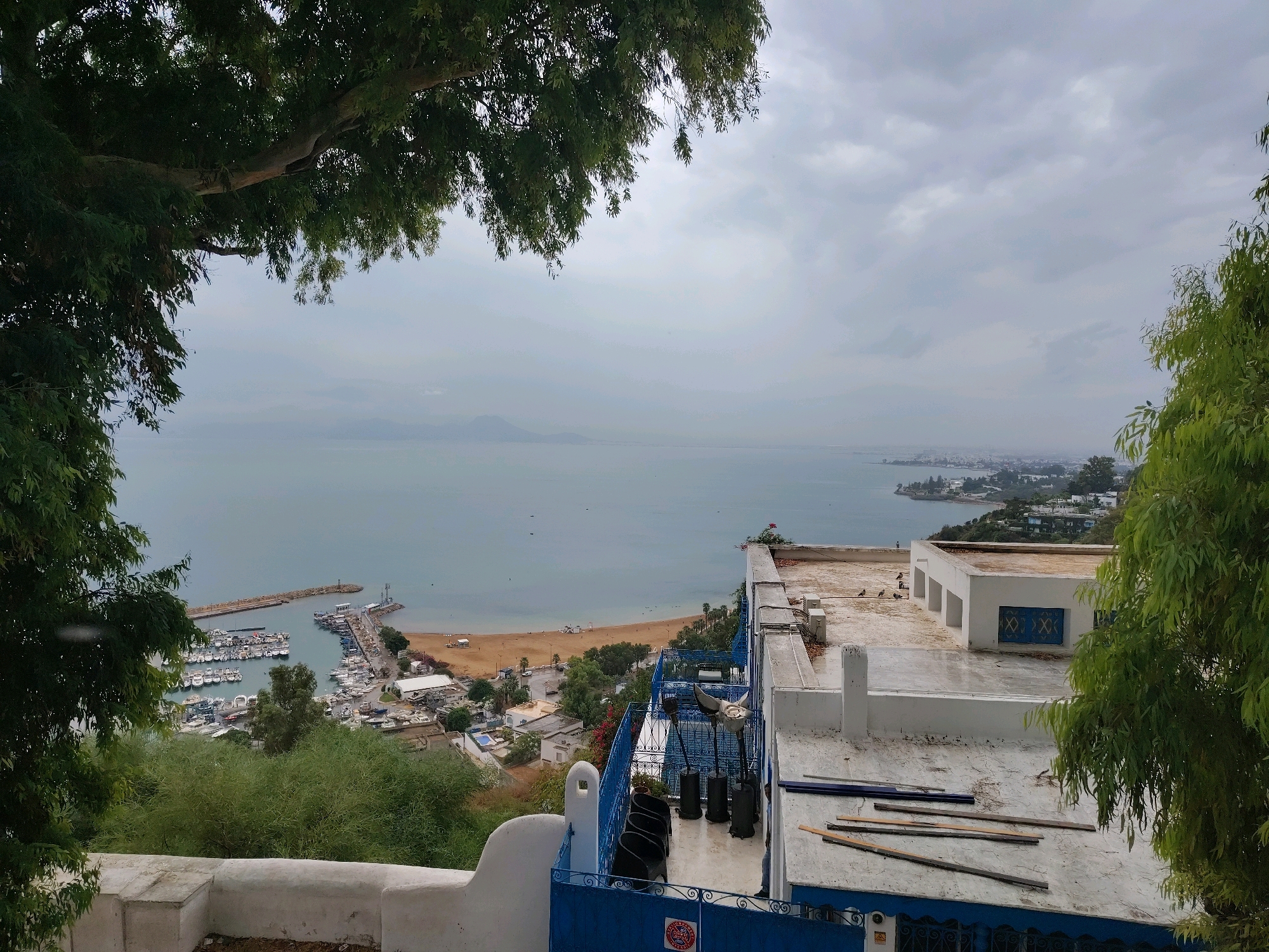 The image depicts a scenic overlook of the city of Tunis, Tunisia, with the Mediterranean Sea in the background. In the foreground, a white stucco wall separates the viewer from a balcony with blue railings and chairs. The balcony offers a view of a harbor filled with numerous boats, a sandy beach, and buildings scattered along the coastline. Lush green trees frame the scene on the left and right sides, adding a natural element to the urban landscape. The sky is overcast, suggesting a cloudy day. The overall impression is one of a coastal city with a bustling harbor and a clear view of the sea.