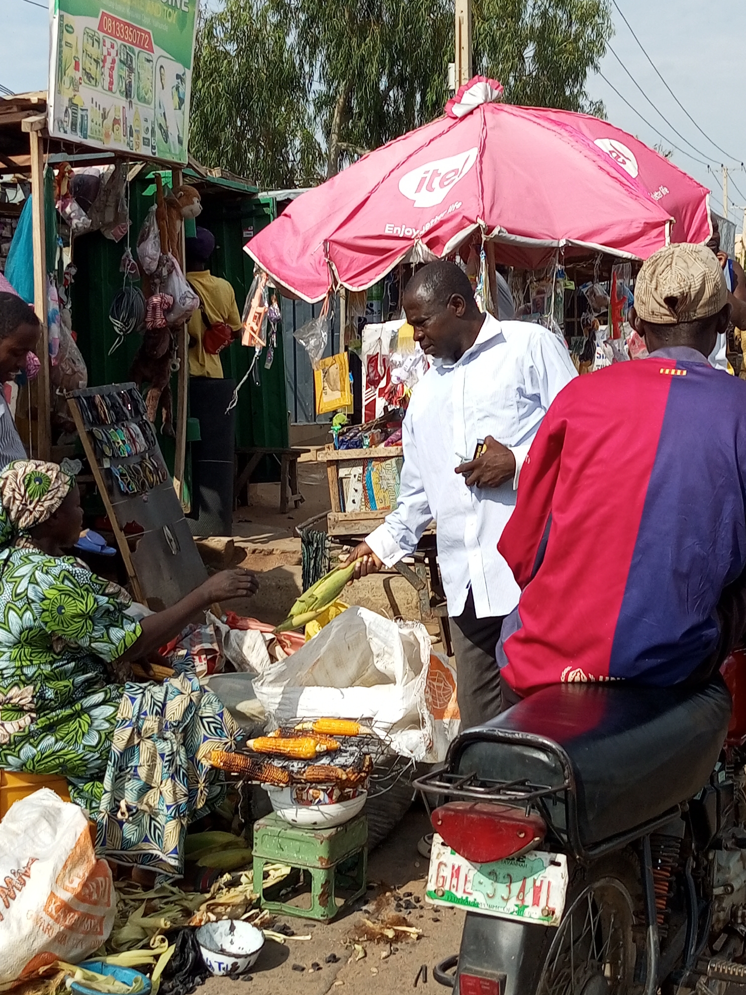 The image depicts an outdoor market scene in Tempure, Nigeria. In the foreground, a woman wearing a green and white patterned headwrap and dress is interacting with a man in a light blue button-down shirt. She appears to be offering him corn. To the right, another person is seated on a motorcycle, wearing a red and blue patterned shirt.
Behind the individuals, various market stalls are visible, adorned with a large pink umbrella with the "itel" logo. Goods are displayed for sale, including what appear to be toys, clothing, and food items like grilled corn on a rack. Bags and other market detritus are scattered on the ground. The environment suggests a typical bustling marketplace.