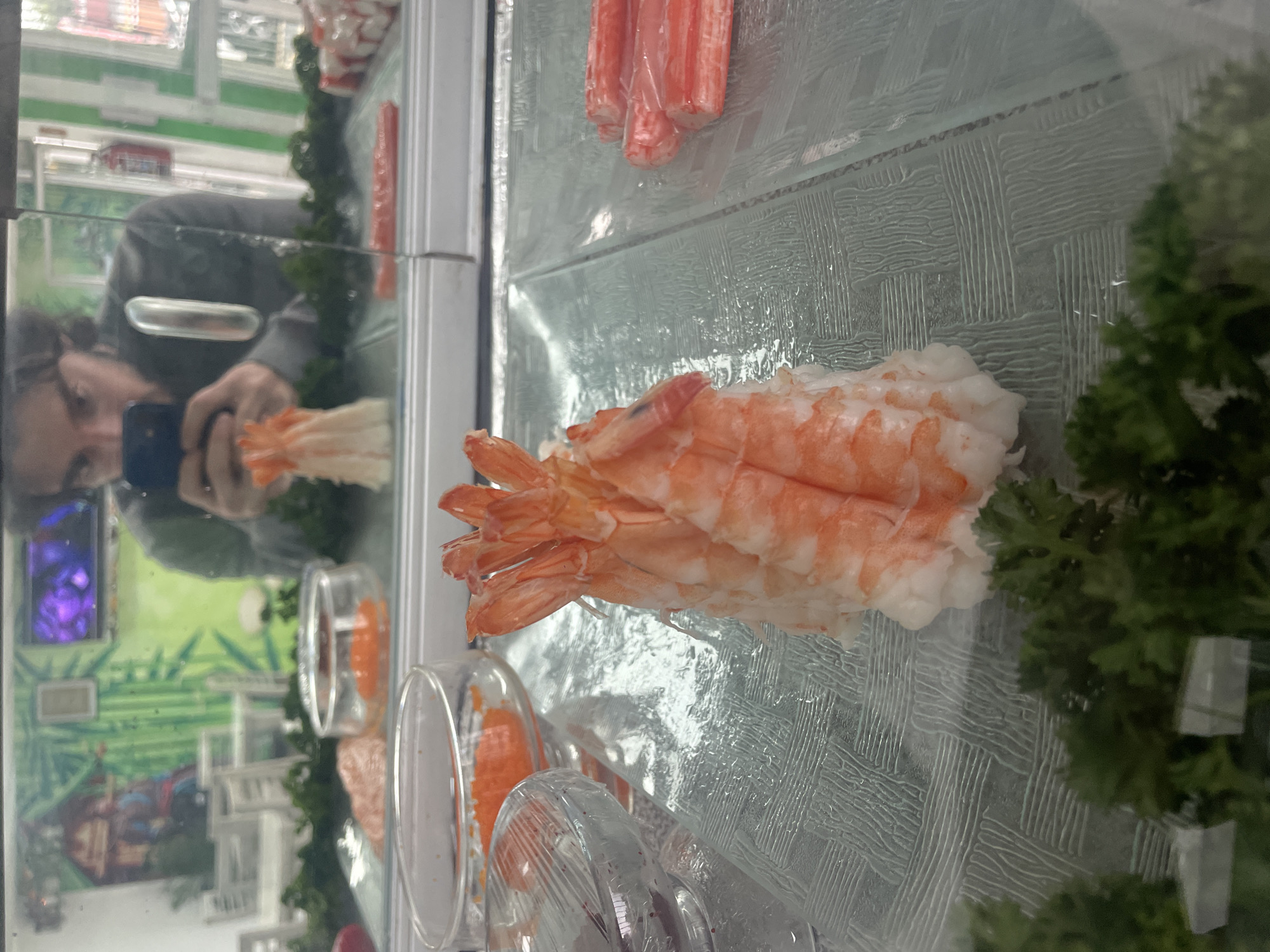 A person is taking a photo of a sushi display case in Naucalpan de Juárez, Mexico. The case contains shrimp, crab sticks, fish roe in small containers, and green parsley. The display case has a textured glass surface. The person is reflected in the glass of the display case, holding a smartphone. The background reflection shows greenery, possibly from a garden or park.