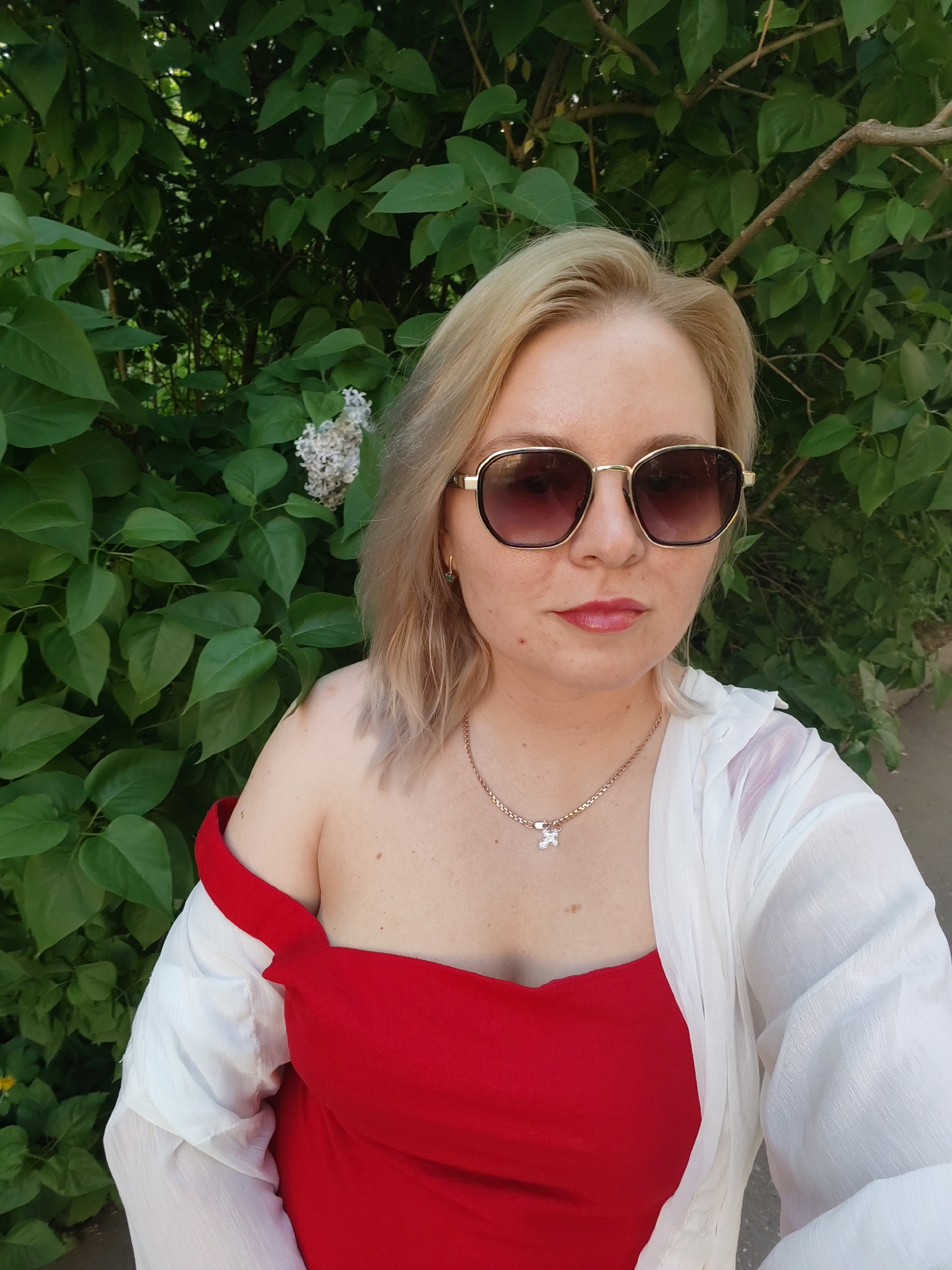 A woman is standing outdoors, likely on a sidewalk or path, with a dense green bush behind her. She is wearing dark sunglasses, a red top, and a white sheer cover-up. She has blonde hair and a gold necklace with a flower-like pendant. There is a small bunch of white flowers on the bush behind her. The photograph was taken in the city of Izhevsk, Russia.