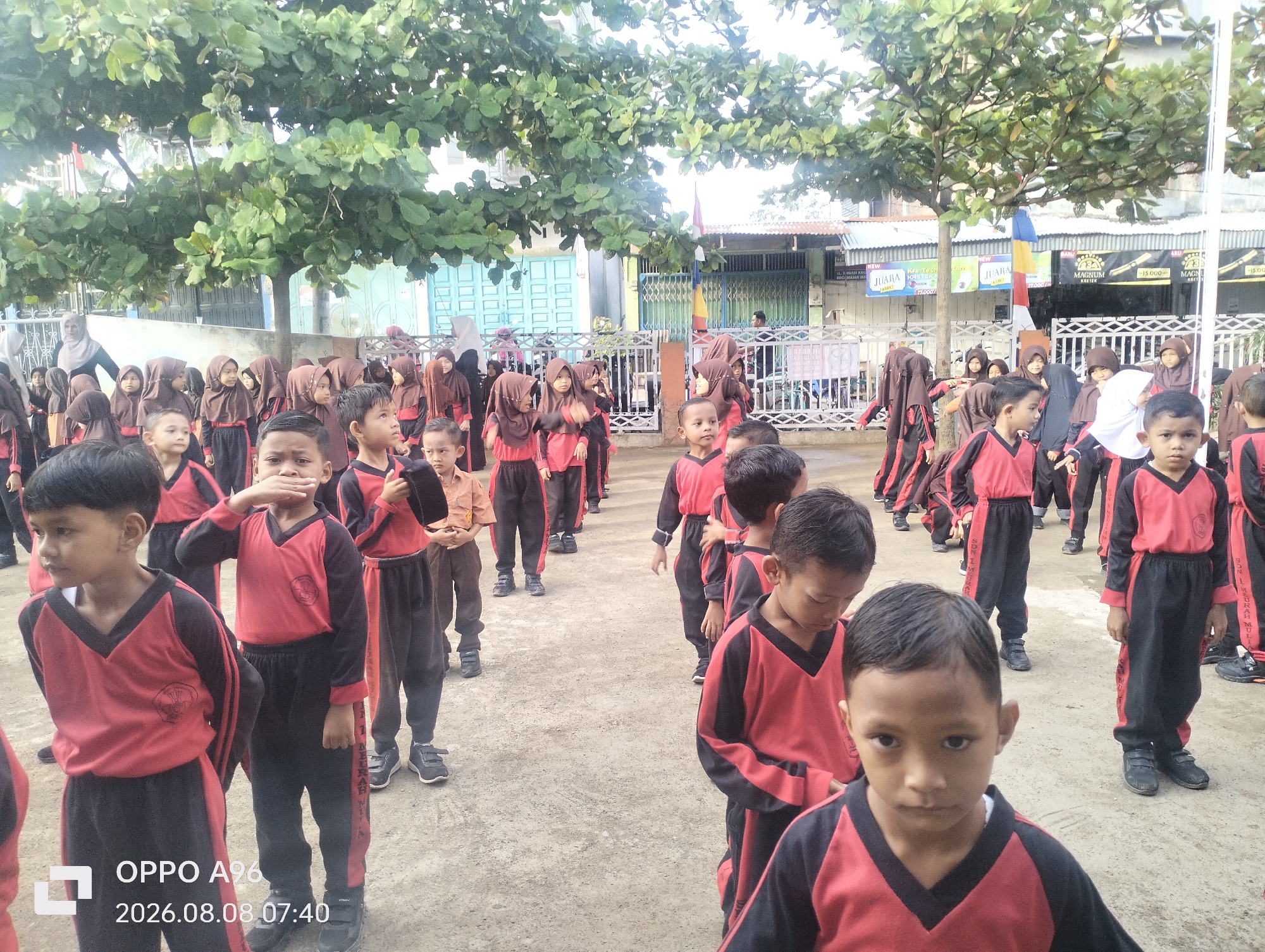 A group of elementary school-aged children are standing in an outdoor schoolyard in Keudekarieng, Indonesia. The children are dressed in matching black and red uniforms with the school logo on the left breast. The schoolyard is paved with gravel and dirt, and there are trees and buildings in the background. Some of the children are wearing headscarves, indicating they are likely Muslim. The children appear to be in a line or formation, possibly for an activity or assembly. One child in the foreground is looking directly at the camera. The timestamp on the image indicates it was taken on August 8, 2026, at 7:40 AM.