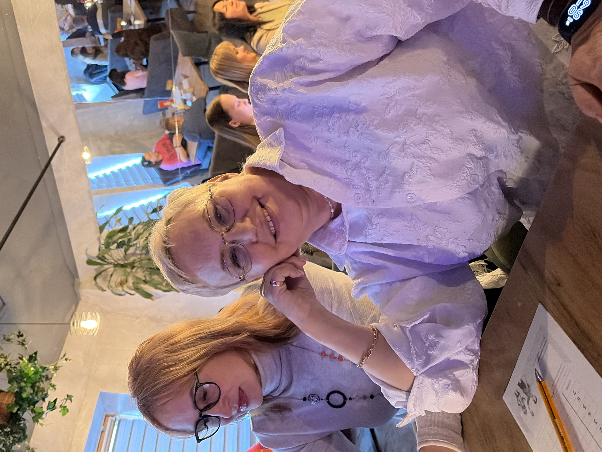 An indoor establishment in Chaikovsky, Russia, resembling a modern cafe or restaurant, is depicted. Two women are seated at a wooden table in the foreground. The woman on the right, with short blonde hair and gold-rimmed eyeglasses, wears a white collared shirt with a textured floral pattern, a pearl necklace, a gold link bracelet on her right wrist, and a ring with a reddish stone on her right ring finger. She smiles directly at the viewer, with her right hand supporting her chin. A dark smartwatch is visible on her left wrist. Behind and to her left, a woman with long blonde hair and black-rimmed eyeglasses wears a light grey turtleneck sweater and two necklaces: one with a black circular pendant, the other with small orange beads. She is looking downwards and to her left, focused on something off-camera. On the wooden table in front of them, a white sheet of paper with numerical markings and a yellow pencil are present. In the background, multiple patrons are seated at other tables, engaged in conversation or personal activities. The environment features modern decor including light-colored walls, exposed ceiling elements, potted plants, hanging artificial ivy, and a mix of warm pendant lighting and cool blue LED strip lighting accentuating horizontal window blinds.
