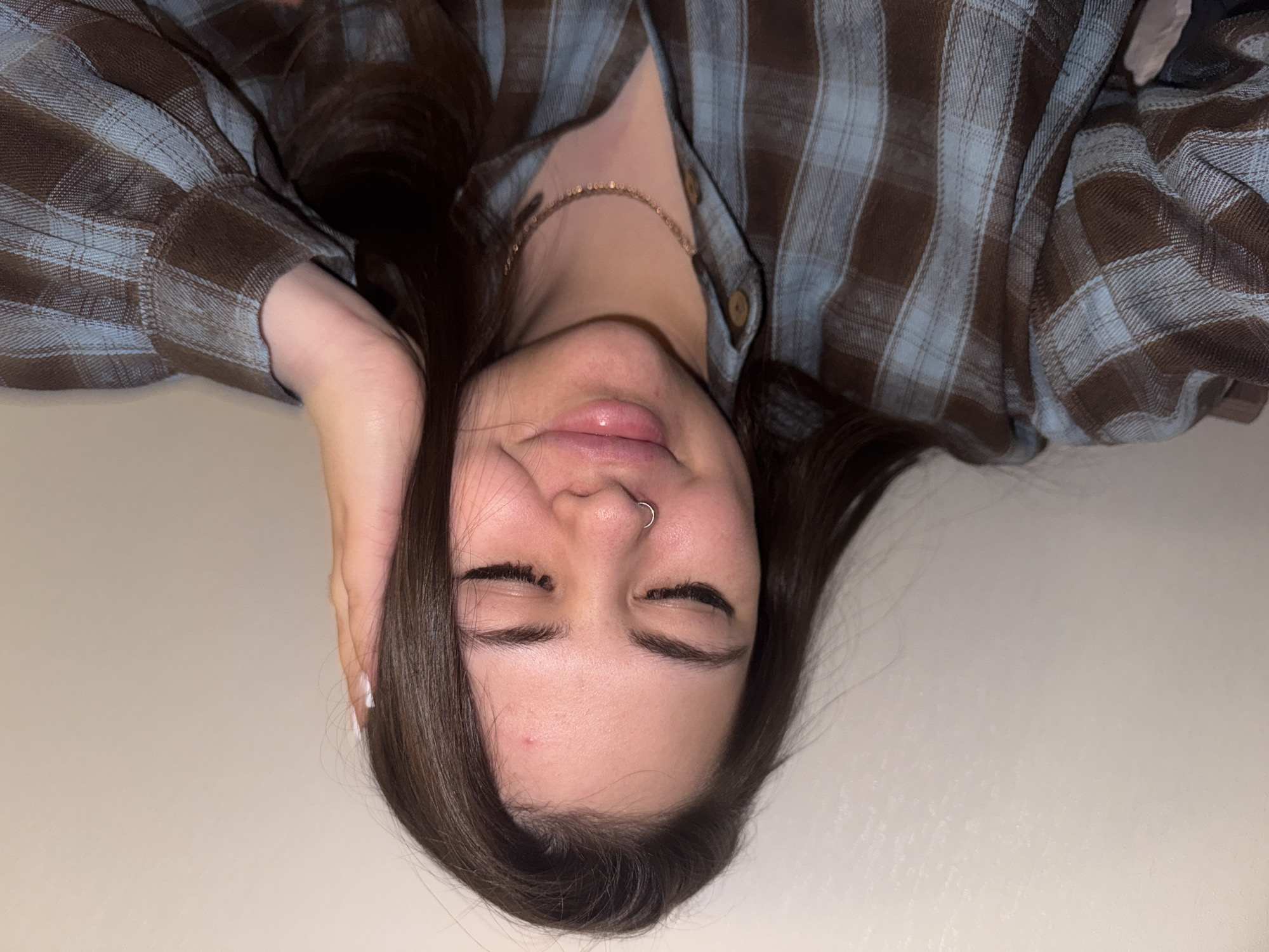 An intimate, portrait-style photograph captures a person, appearing to be a young adult female, resting in an indoor setting in Bugry, Russia. The image is oriented with the person's head positioned at the top. She has long, dark brown hair that frames her face and spreads out around her. Her eyes are closed, suggesting a moment of peace or rest, and a slight, relaxed smile plays on her lips, which are slightly parted. A silver hoop nose ring is visible in her left nostril.
She is wearing a long-sleeved plaid shirt featuring a pattern of brown and light blue checks. A delicate, gold-colored chain necklace adorns her neck. One of her hands is gently placed behind her head or ear, with her fingernails painted white. The background is a plain, light-colored surface, possibly a wall or bedding, indicating a simple and uncluttered environment. The lighting is soft and diffuse, highlighting the textures of her hair and clothing. No text is visible in the image. The overall impression is one of calm and comfort.