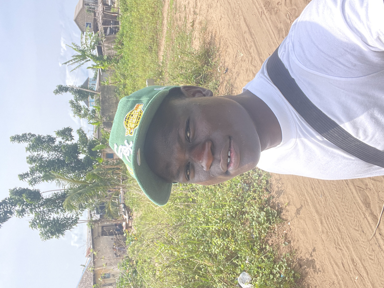 The media file is a portrait-style photograph taken outdoors on a bright, sunny day. The image is oriented sideways, with the subject's head tilted to the right. The central figure is a young person with dark skin, looking directly at the camera with a faint, pleasant smile. They are wearing a plain white t-shirt and a green baseball cap. A black strap, likely from a bag, is visible across their chest and shoulder.
The green baseball cap features a prominent yellow and white embroidered logo on the front. The text "WORLD SERIES" is clearly visible in yellow lettering, surrounded by a circular emblem in yellow and orange tones.
The background depicts an outdoor setting characterized by a sandy or dirt path to the right of the subject and lush green vegetation and grass covering the left and mid-ground. Several trees, including a prominent palm tree and other leafy varieties, are visible throughout the landscape. In the distance, several residential buildings or houses with simple architecture can be seen under a clear, bright sky. The strong natural light suggests it is midday or early afternoon. A small piece of white plastic litter is discernible on the ground among the vegetation. The scene portrays an everyday environment in a semi-rural or developing residential area, consistent with the provided location context of Badagry, Nigeria.