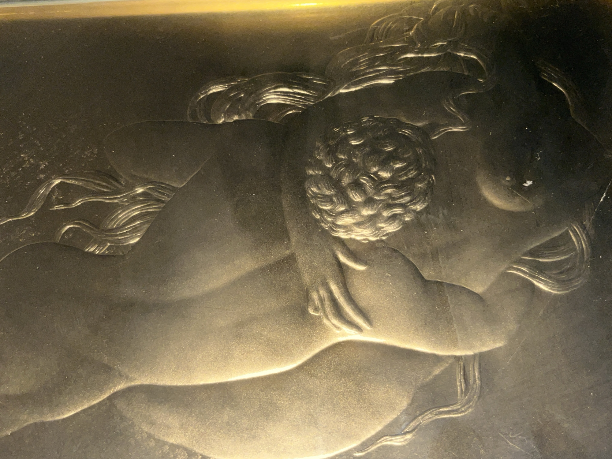 The image depicts a bas-relief sculpture of a woman and child. The figures are nude and rendered in a style reminiscent of classical art. The woman is reclining with her hair cascading around her head. The child is nestled against her chest, their arms wrapped around each other. The sculpture appears to be made of a metallic material, possibly bronze or copper, and has a slightly tarnished or aged appearance. The image does not provide sufficient information to determine its location in the city of Tinaquillo, Venezuela.