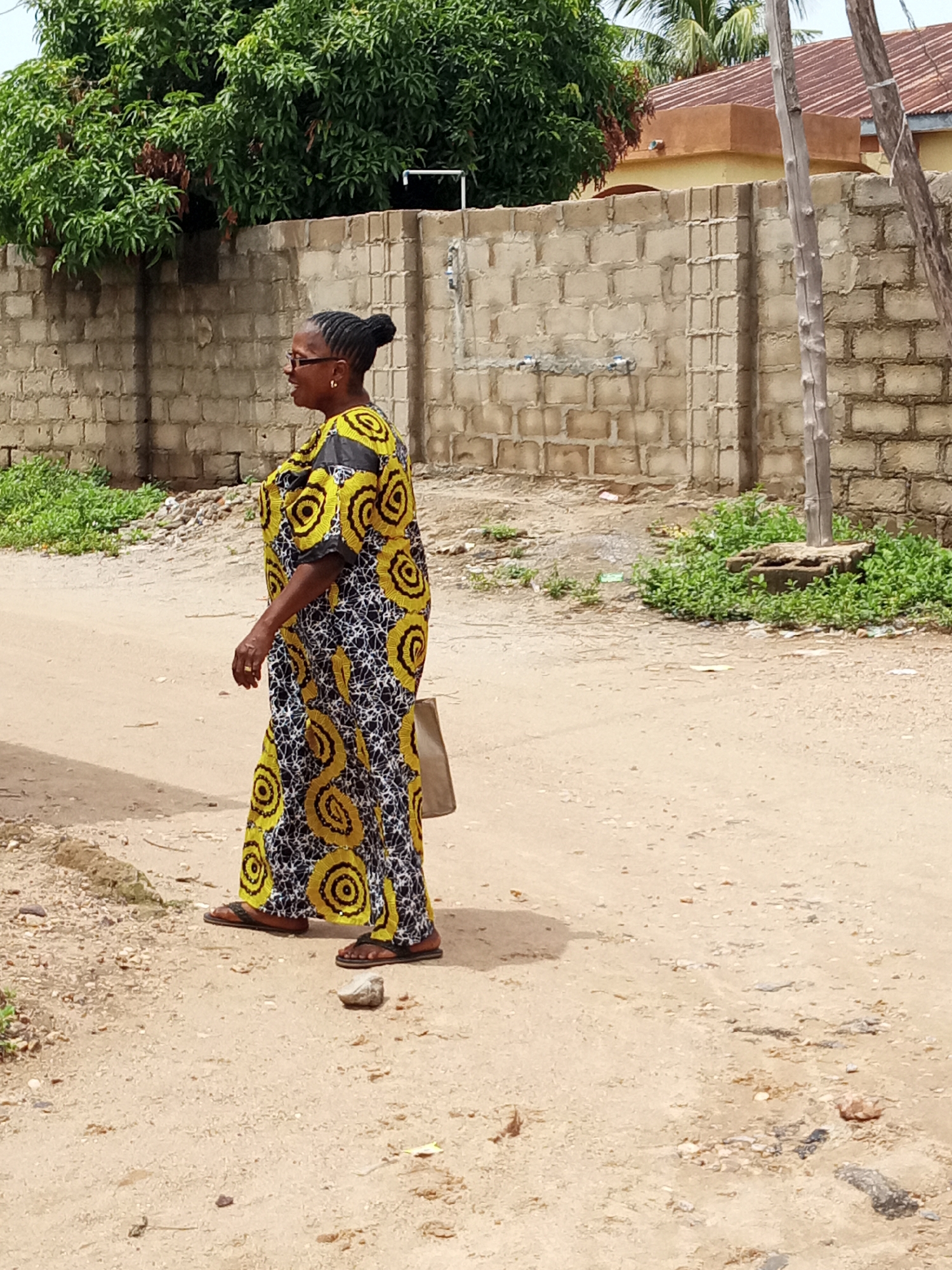 A medium shot captures a mature woman in a black, white, and yellow patterned dress, standing on a dirt road in an urban area of Unknown, Unknown. She is turned to her left, wearing glasses, and her hair is styled in a bun. Her clothing is a floor length dress with bold yellow circle patterns within the black and white print. She is also wearing sandals. A beige bag hangs from her left shoulder.
The backdrop includes a weathered concrete block wall, green foliage, and a glimpse of a tan colored building. The area appears to be a residential neighborhood with a dirt road surface. The lighting suggests it is daytime.