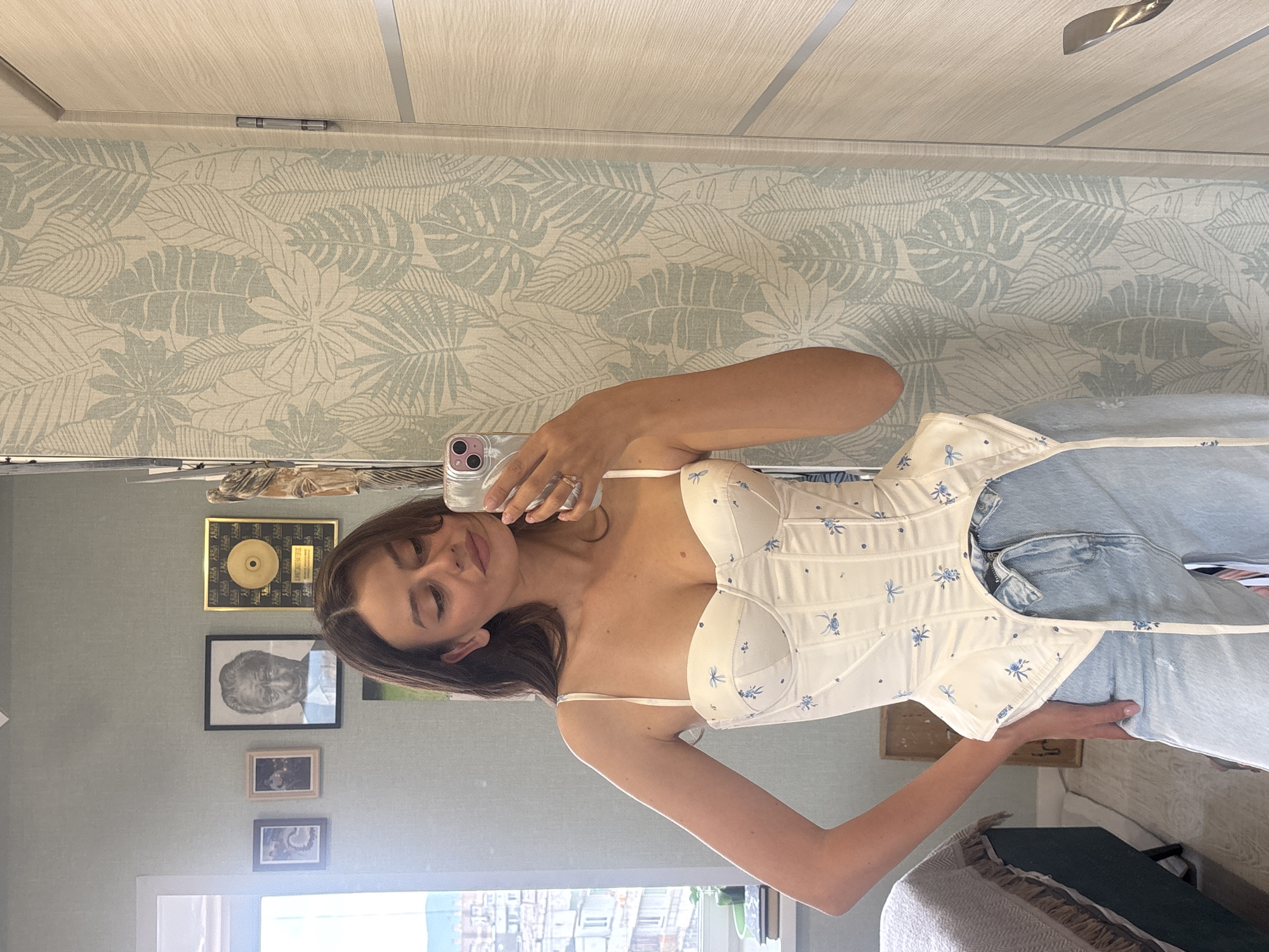 A young woman with long, brown hair takes a mirror selfie in a room. She is wearing a white corset with a blue floral pattern and light blue jeans. She is holding a silver smartphone in her right hand, which is angled to capture her reflection. Behind her, the wall is covered in a light green and white tropical leaf wallpaper. To the right of the wallpaper is a wooden door. To the left of the woman, a window reveals a cityscape with buildings and a cloudy sky, suggesting an urban environment. On the wall behind her, above the window, are several framed pictures and an award. A bed with a dark blue cover is partially visible to the far left. The scene appears to be an indoor residential setting.
