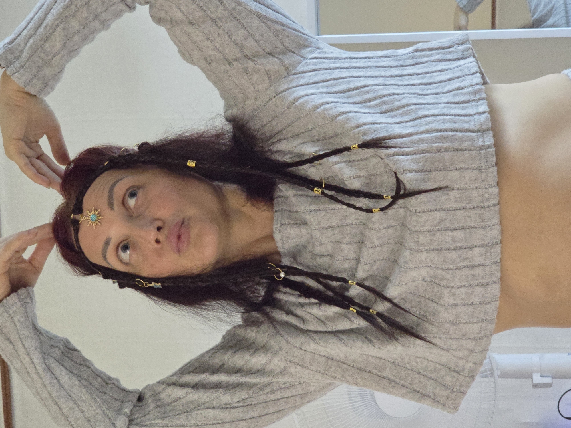 A woman with dark hair, adorned with a golden sun-shaped hairpiece with a turquoise gem and several smaller dangling charms, is positioned centrally in the frame. She is wearing a grey, ribbed, cropped sweater. Her hands are raised and positioned above her head, as if forming bunny ears. Her gaze is directed upwards and to her left. Her hair is partially braided and decorated with small golden rings and beads. The background consists of a plain, off-white wall. To the left of the woman, a white oscillating fan is partially visible. The overall impression is a close-up, indoor shot with a focus on the woman's attire and adornments. The location in the city of Pyeongtaek-si, South Korea is not discernible from the image.