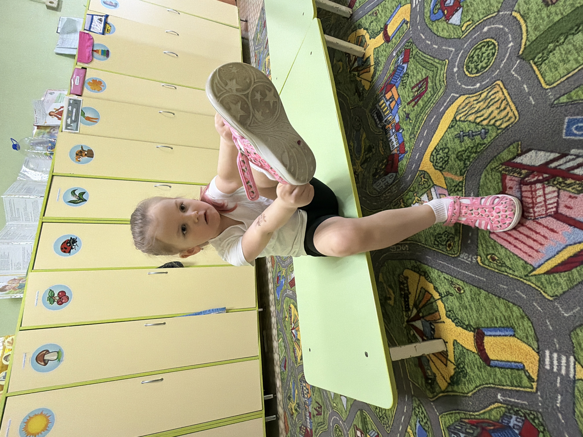 A young girl is sitting on a green table in a room, holding up a pink shoe with a white sole to her face. She is wearing a white t-shirt, black shorts, and a matching pink shoe on her right foot. The room is lined with yellow lockers featuring pictures on them. A rug with a colorful street map design covers the floor. The location is Chaikovsky, Russia.