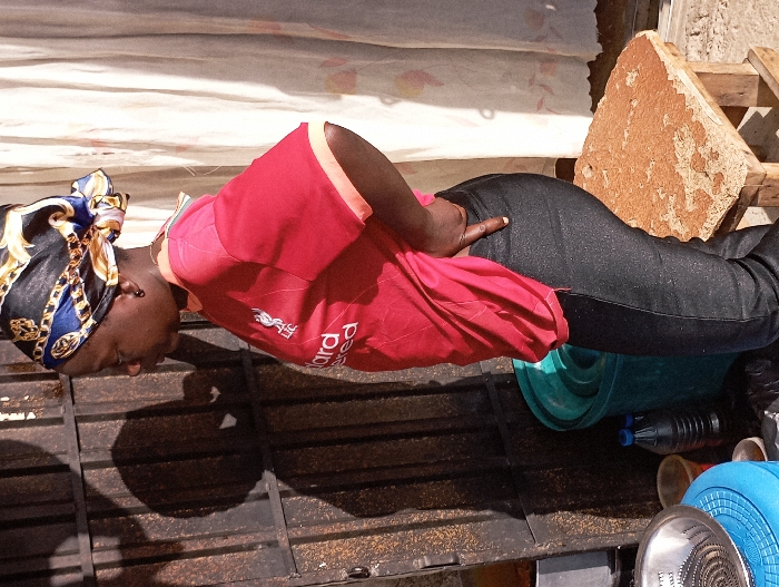 This image, captured in Jos, Nigeria, depicts an adult female standing in a bright outdoor or semi-enclosed setting. The subject, viewed from her left side, has dark skin and wears a black, blue, and gold patterned headscarf and small dark stud earrings. She is dressed in a red short-sleeved t-shirt, identified as a Liverpool FC jersey by the visible "LFC" crest and "Standard Chartered" sponsor logo, paired with dark, form-fitting trousers. Her right hand is positioned on her hip, with the index finger extended downwards.
In the foreground, to the subject's left, a light blue plastic stool supports a silver metallic colander with a spiral pattern. Directly behind the stool and partially obscured by the subject's leg, a dark green plastic bucket is present. On the ground nearby are two plastic bottles—one clear with dark liquid, one opaque—and a small orange container. To the subject's right, a small wooden stool with a rough, irregular, reddish-brown top surface, consistent with stone or compacted earth, is visible.
The background on the left features a dark metallic door or gate with vertical panels and visible signs of wear, upon which a distinct human head and shoulder shadow is cast. On the right, a light-colored curtain or fabric is partially visible. Strong shadows indicate direct sunlight.
