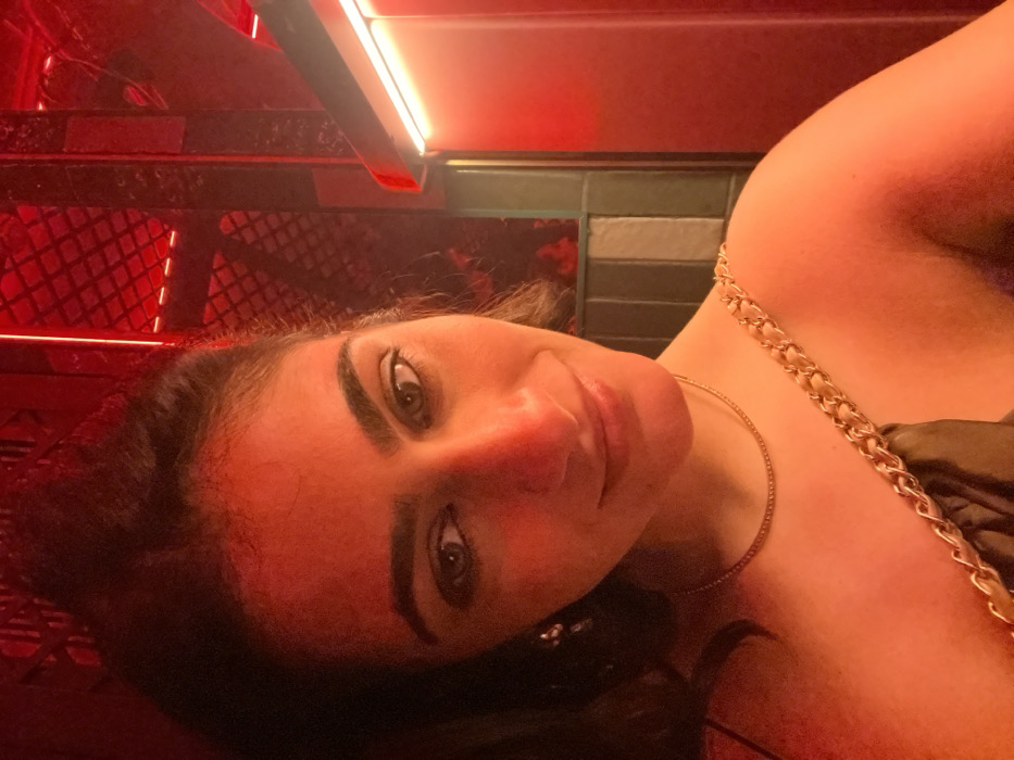 A close-up, chest-up portrait features a single female individual looking directly into the camera with a slight smile. She has dark hair, parted centrally, dark eyes accentuated with eyeliner, and dark, defined eyebrows. A small, light-colored dangling earring is visible on her left ear. She wears a thin, golden chain necklace and a dark top with a prominent gold chain strap over her right shoulder. Her skin tone appears warm, heavily influenced by the ambient red lighting. The background is dimly lit and saturated with a strong red hue, indicating an indoor setting such as a bar or club. Visible elements in the background include a dark, grid-like structure in the upper right, vertical panels or columns below it (some appearing dark or with a subtle greenish-grey striped pattern under the red light), and a horizontal bright red linear light source along the far right edge of the frame. The location in the city of Madrid, Spain, as specified, cannot be independently confirmed through visual elements within the image itself, which solely depicts an interior space.