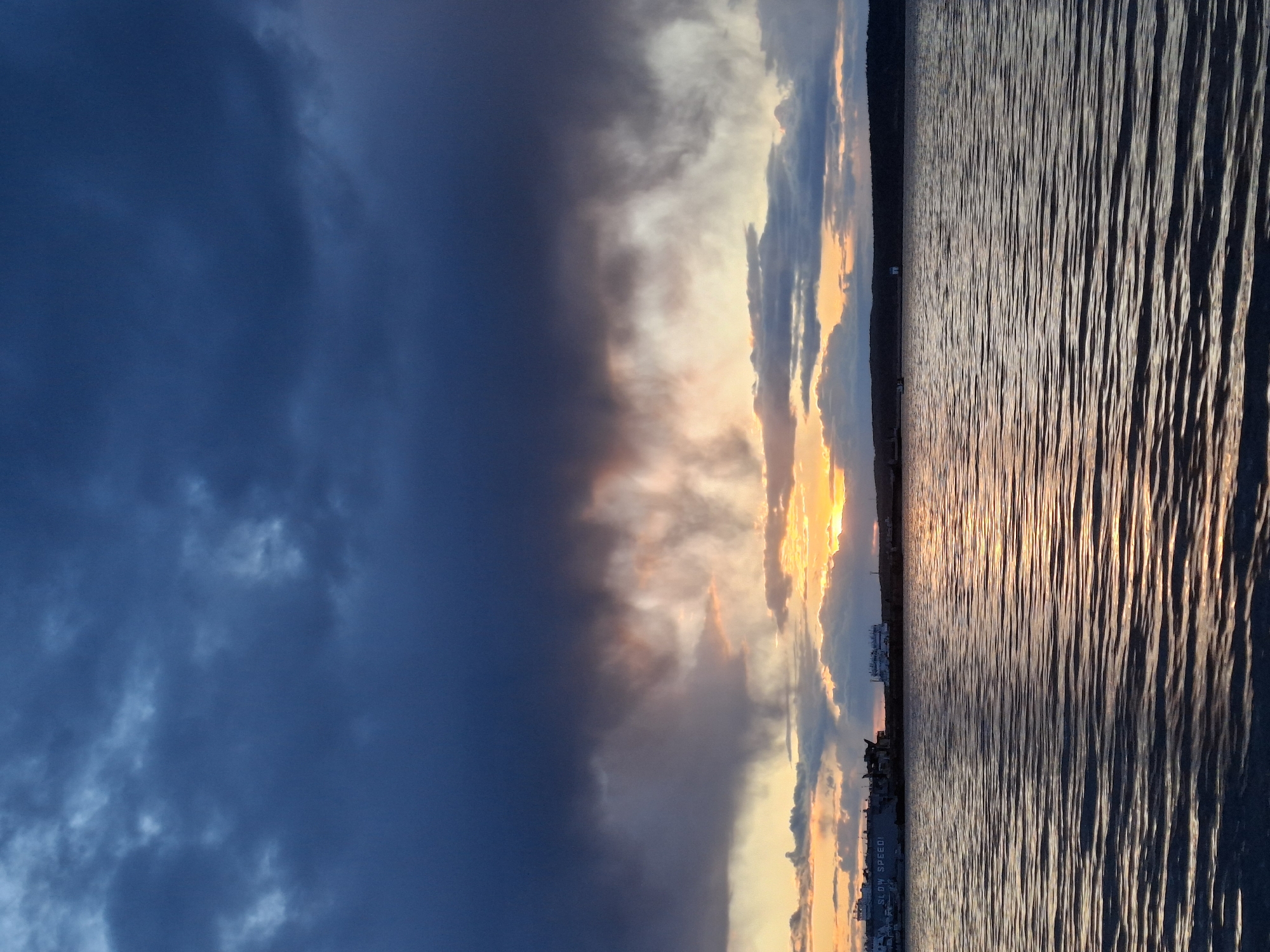 A slightly tilted, landscape-oriented photograph shows the skyline and water in the city of Chaikovsky, Russia, at twilight. On the left of the image, dark, brooding clouds fill about half the frame, with a hint of lighter clouds interspersed. To the right, the clouds transition to lighter and more colorful hues of orange, yellow, and gray as they approach the horizon. The water in the foreground, and on the right side of the image, reflects the colors of the sky, with a shimmering, rippled surface. In the middle ground, near the horizon line, there are silhouettes of buildings and structures, giving a glimpse of the city. The photograph captures the contrast between the dark clouds and the illuminated water and sky. The absence of any visible people or animals emphasizes the vastness and serenity of the location at twilight.