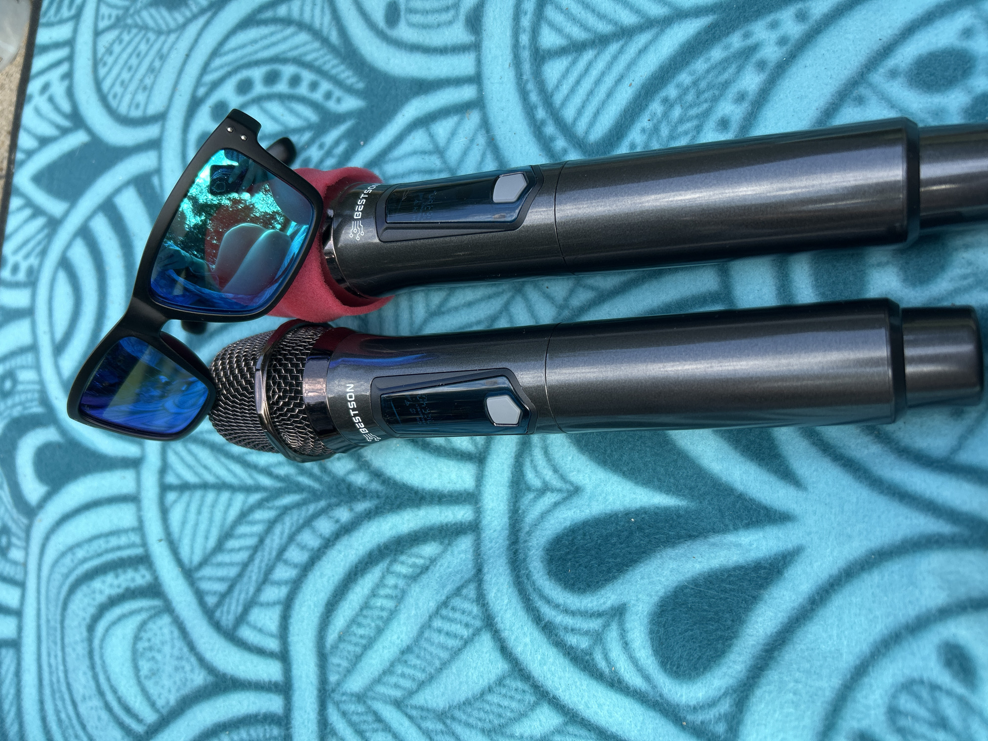 Two dark gray microphones are placed on a turquoise towel with a circular pattern. One microphone has a red foam windscreen on its head. A pair of black sunglasses with blue mirrored lenses rests on top of the microphones. The sunglasses reflect a person holding a camera. The scene is outdoors, as indicated by the bright lighting and reflections. The exact location is not specified in the image or its context.
