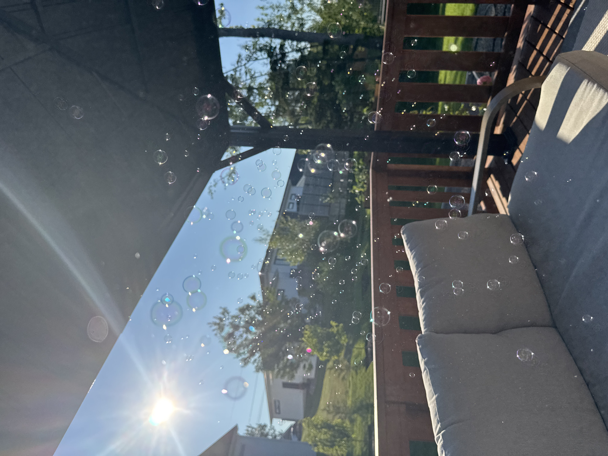 This image depicts a sunny outdoor scene, likely a backyard deck, with numerous bubbles floating in the air. The foreground features a portion of an outdoor sofa with beige cushions, casting a shadow from sunlight filtering through an overhead structure. A wooden railing is visible behind the sofa, and beyond that, a green lawn and trees are seen. In the background, houses and more trees are visible under a clear blue sky. The sun is prominent in the upper left quadrant, with lens flare and light rays extending outwards. The bubbles are scattered throughout the frame, some appearing iridescent. There are no people or animals visible in the image. The location in Dieppe, Canada, is not explicitly indicated in the image itself.