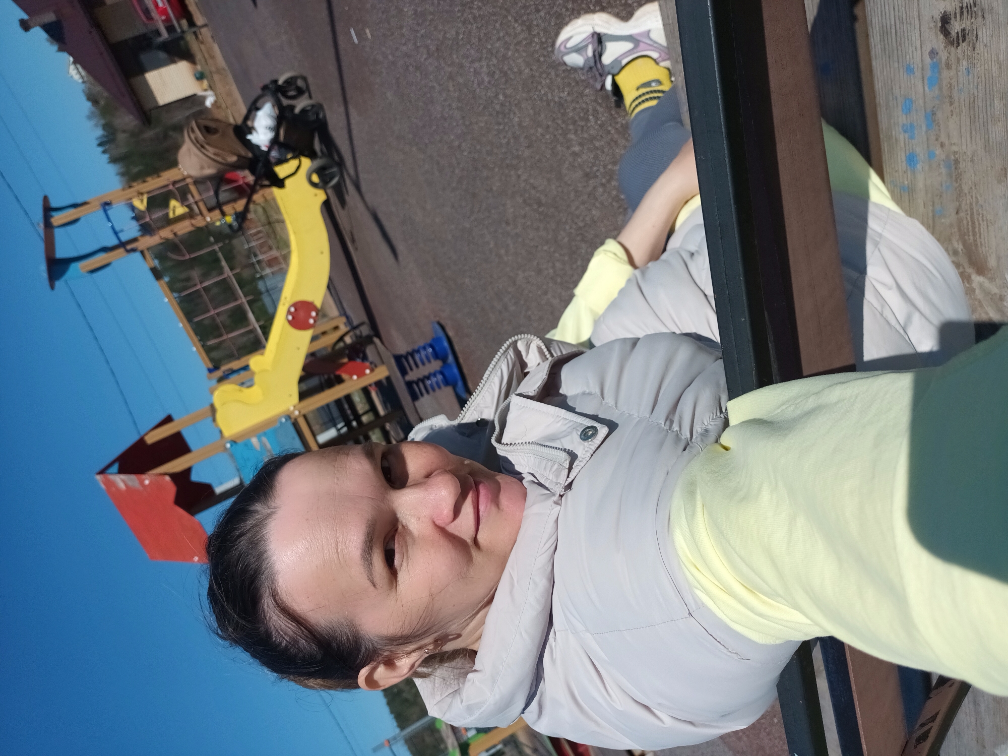 A woman is lying on a park bench in Ольховка, Russia. The woman, who appears to be middle-aged, is wearing a light beige puffer jacket over a pale yellow shirt, gray leggings, and white sneakers with yellow socks. The playground is visible in the background, with a yellow slide, climbing structure, and other play equipment. A stroller is also in the vicinity of the playground. The sky is clear and blue, indicating it's likely daytime. The photo appears to be a selfie taken by the woman herself.